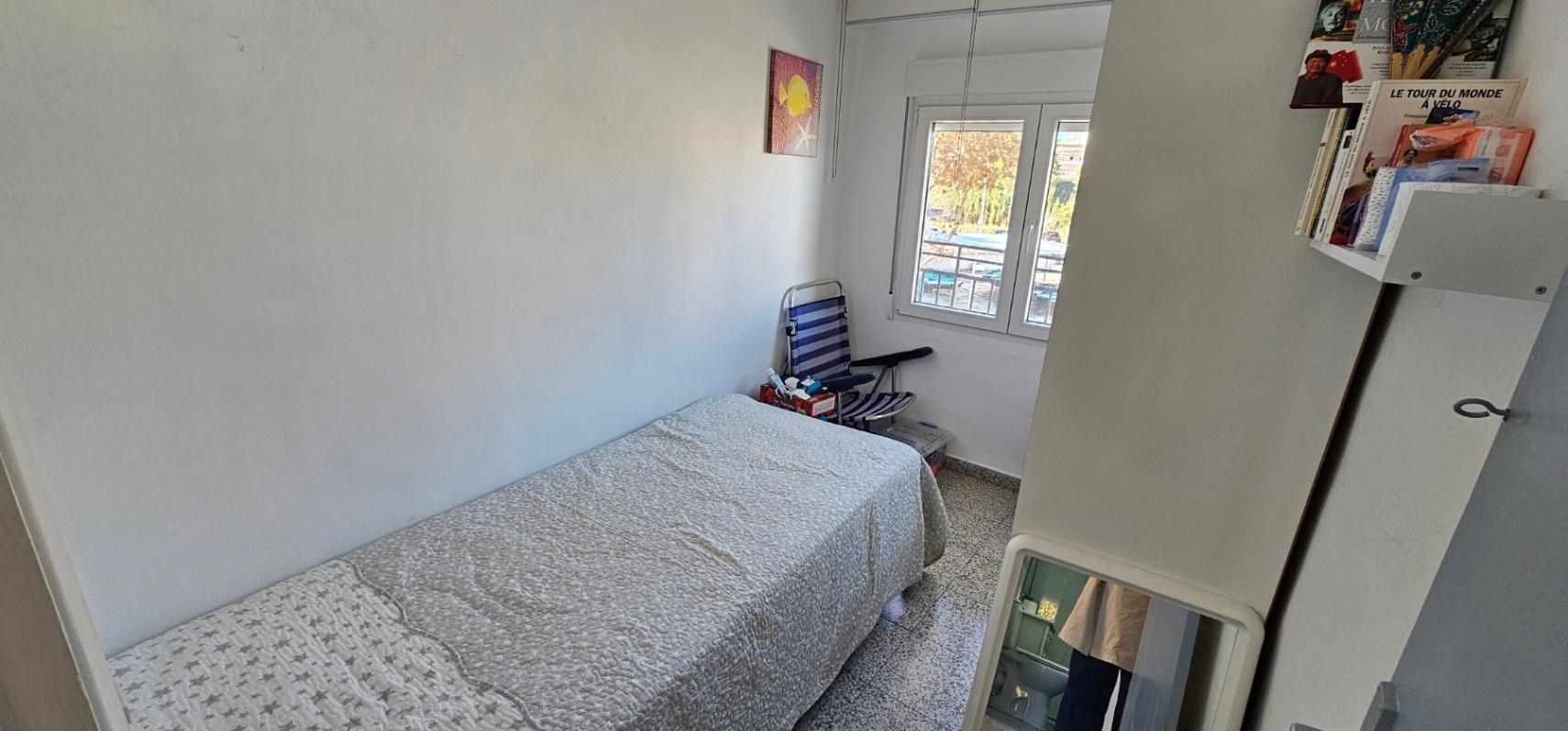 Chambre individuelle avec lit simple, sol en terrazzo d'origine et fenêtre offrant une lumière naturelle. Les murs blancs et les étagères optimisent l'espace.