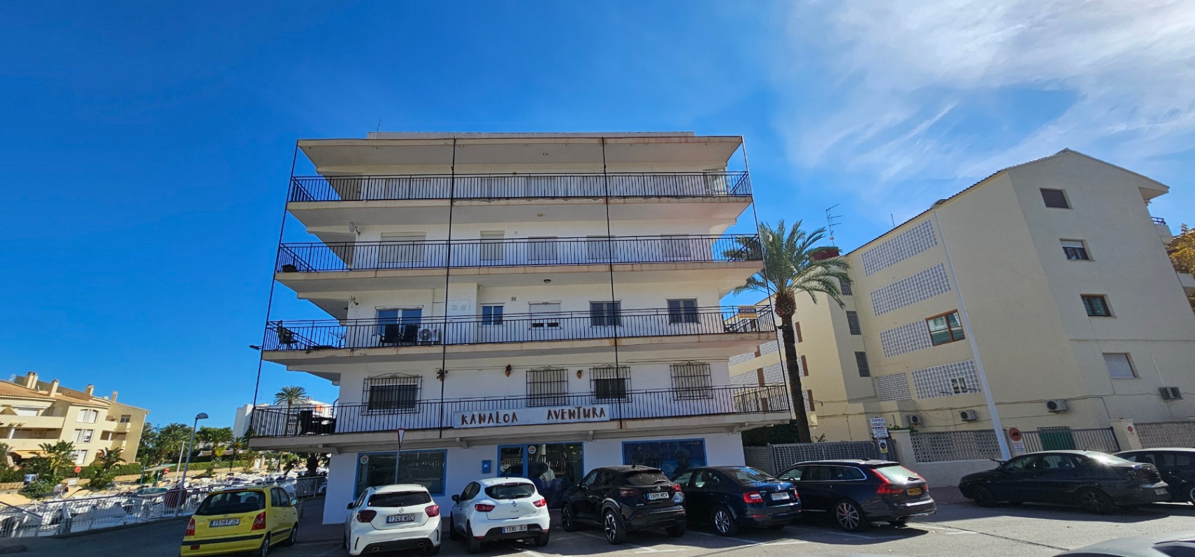 Façade extérieure du bâtiment de style méditerranéen avec de larges terrasses enveloppantes et des garde-corps en métal noir, à proximité du parking.