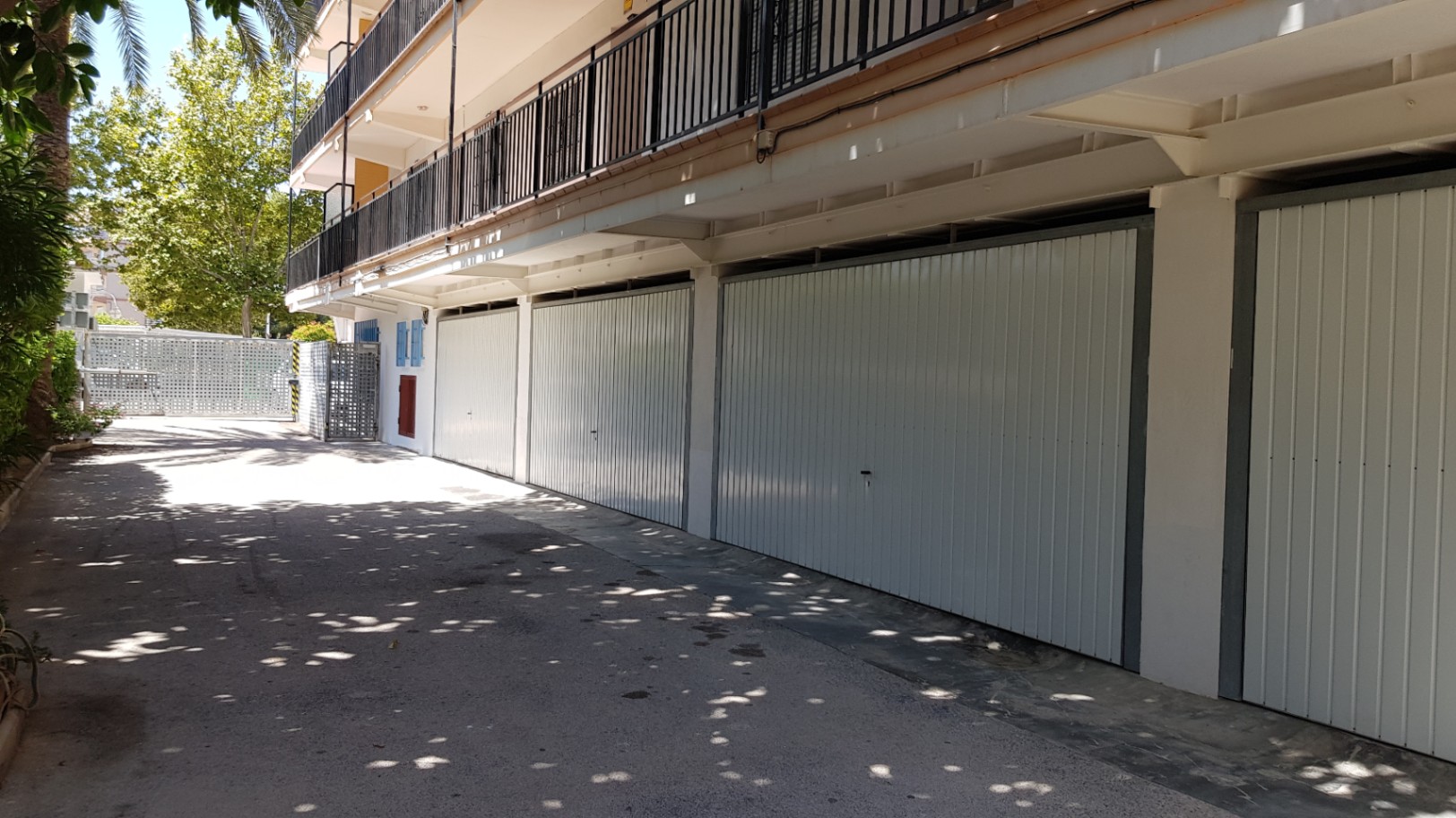 Garages privés individuels avec portes métalliques blanches, situés au rez-de-chaussée de l'immeuble avec un accès direct depuis l'allée pavée et ombragée.