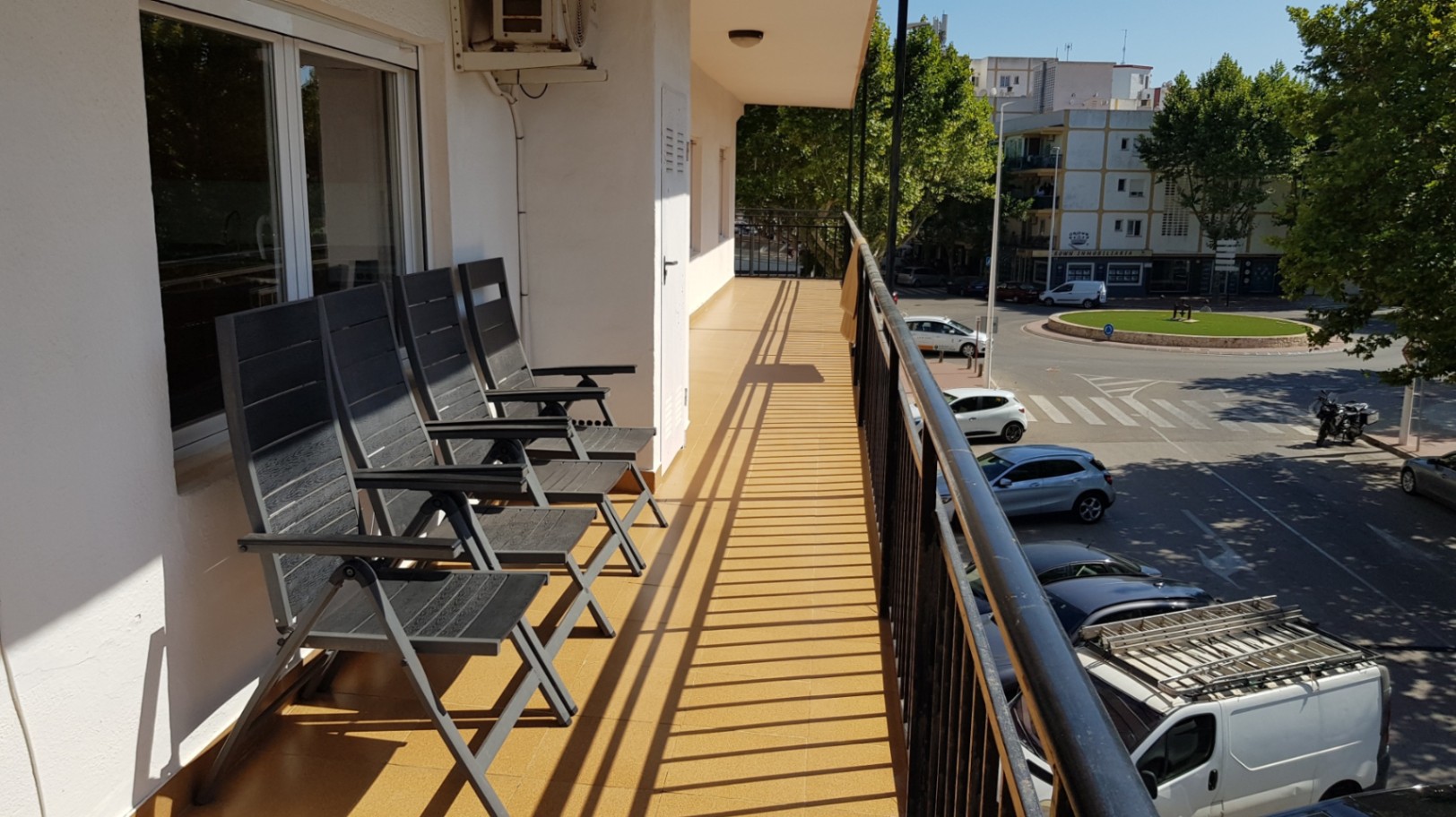 Long balcon avec carrelage ocre, chaises pliantes grises et vue sur le quartier urbain environnant et la végétation locale.