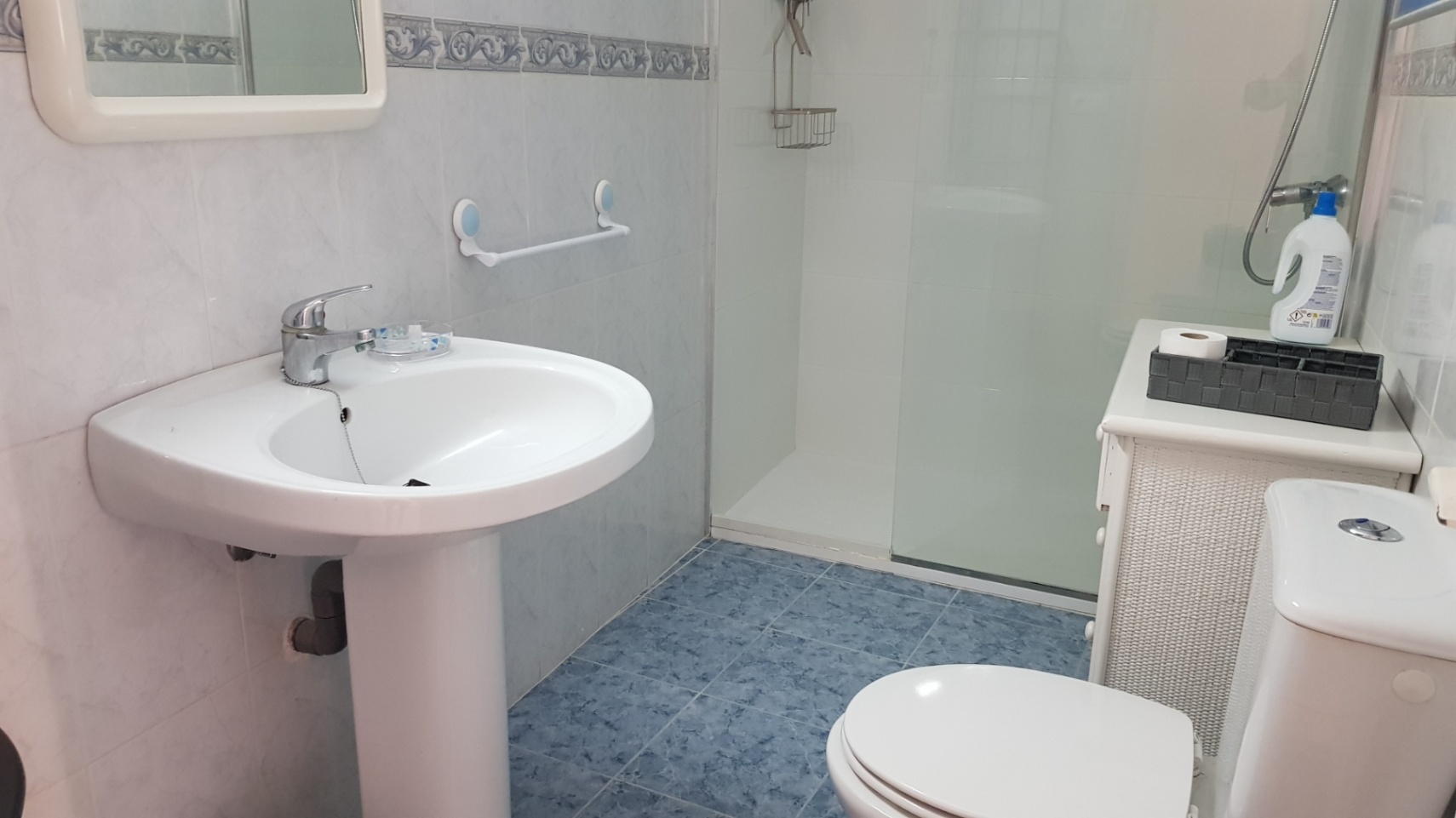 Salle de bain avec carrelage bleu, lavabo sur colonne et douche avec paroi vitrée. Murs carrelés de blanc avec frise décorative et meuble de rangement.