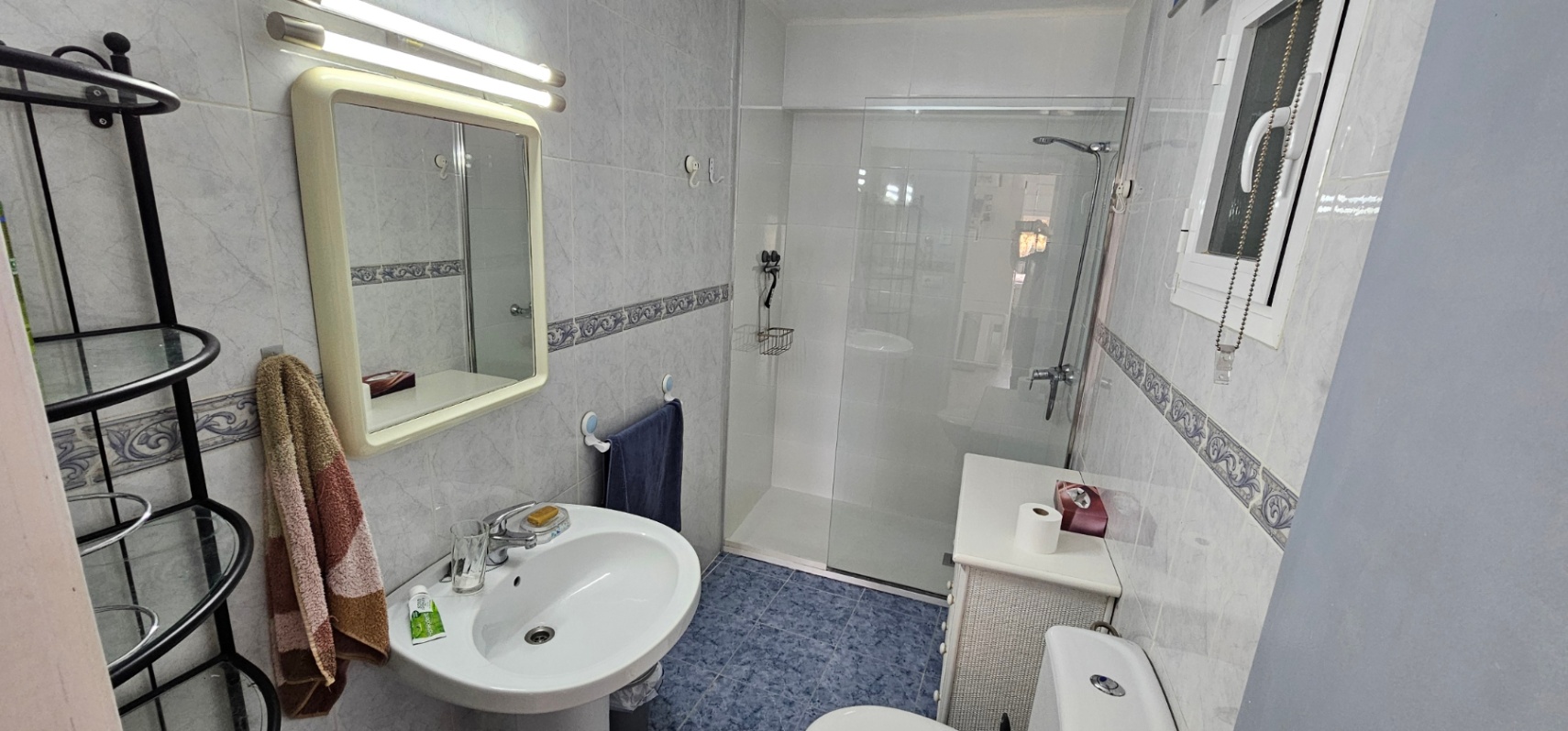 Salle de bain fonctionnelle avec douche à l'italienne, paroi vitrée, lavabo sur colonne et carrelage au sol bleu contrastant avec les murs clairs.