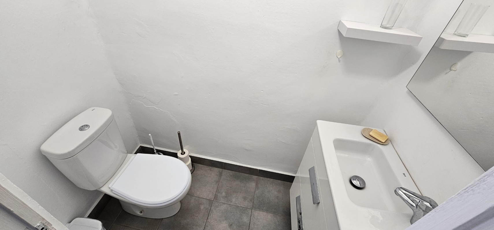 Salle de bain fonctionnelle avec toilettes blanches, meuble vasque avec rangement et carrelage gris au sol. Elle dispose d'un miroir et d'une petite étagère murale.