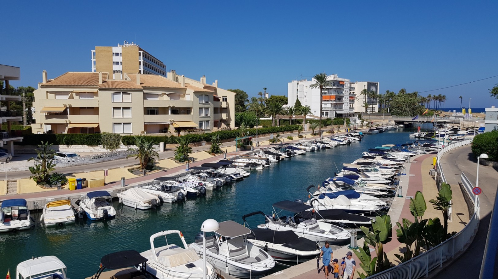 Vue panoramique depuis la terrasse sur le canal de la marina, avec des bateaux de plaisance amarrés et des bâtiments résidentiels sous un ciel dégagé à Jávea.