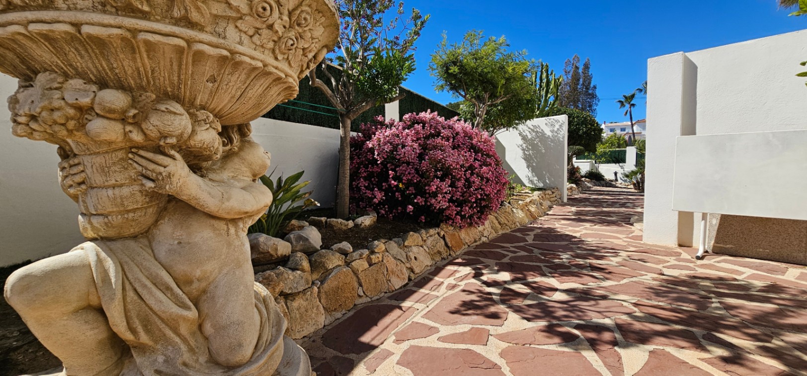 Allée en pierre naturelle bordée d'une statue ornementale et d'une végétation méditerranéenne luxuriante avec des lauriers-roses en fleurs.