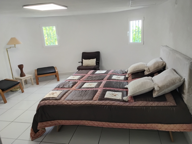 Chambre avec sol en carrelage blanc, lit double et fauteuils d'appoint. Elle dispose de fenêtres avec grilles de sécurité et d'un éclairage par panneau LED intégré au plafond.
