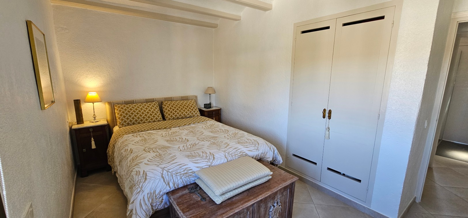 Chambre chaleureuse avec poutres apparentes au plafond, placard encastré blanc et un coffre en bois rustique au pied du lit double.