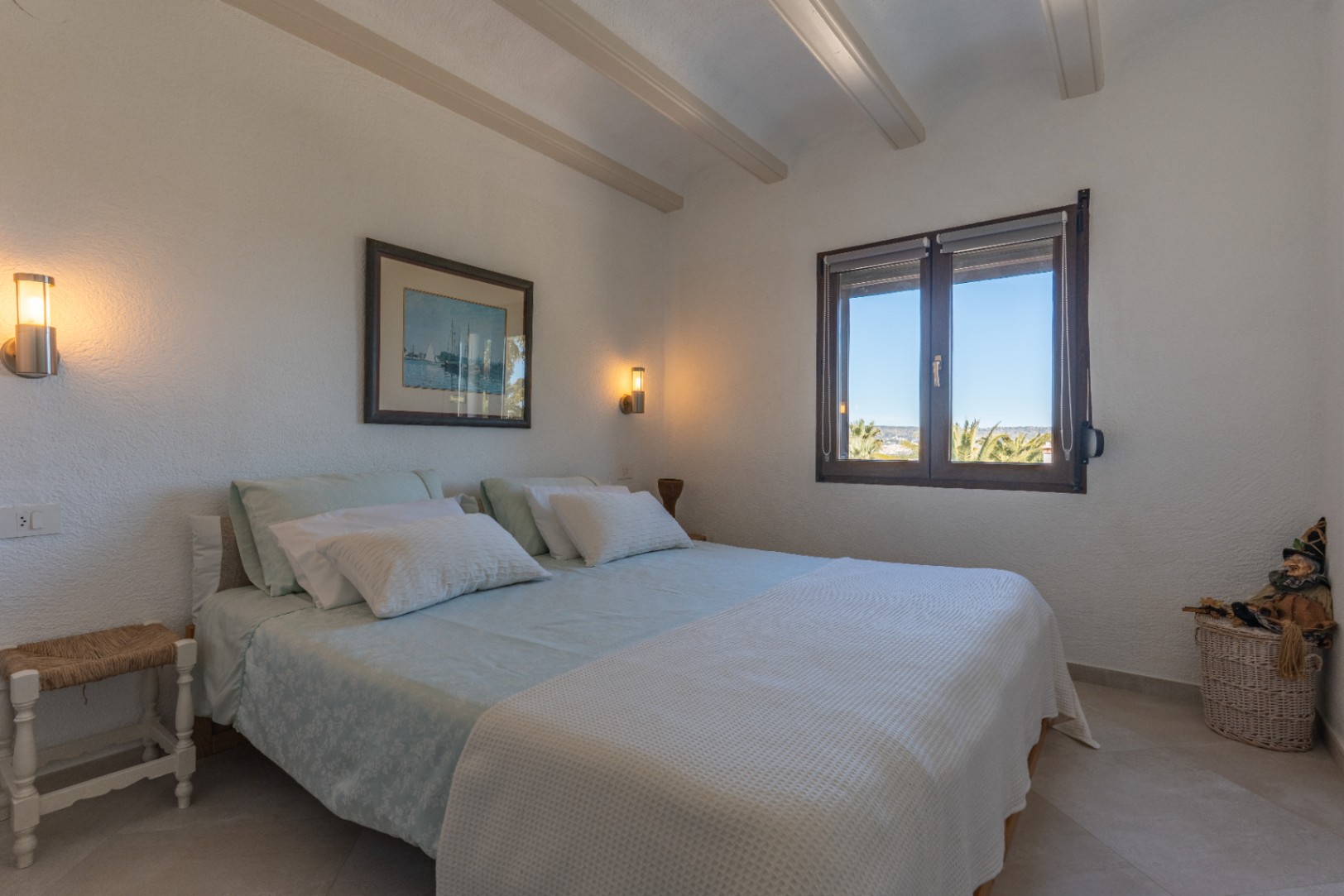 Chambre lumineuse avec poutres apparentes décoratives, appliques murales modernes et fenêtre en bois offrant une vue dégagée sur les palmiers.