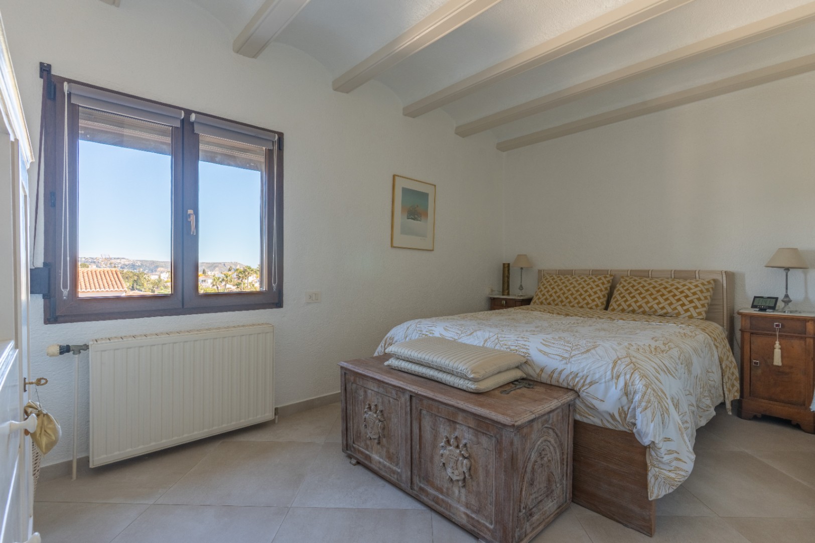 Chambre lumineuse avec poutres apparentes, fenêtre en bois offrant une vue dégagée et un coffre en bois sculpté au pied du lit double.