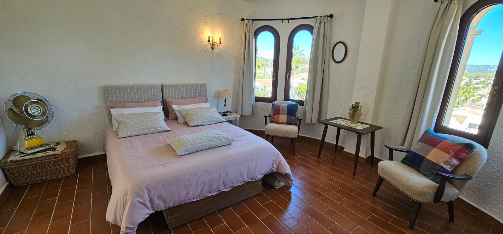 Chambre lumineuse avec sol en terre cuite, fenêtres cintrées en bois et mobilier rustique. Elle dispose d'un coin salon avec fauteuils offrant une vue dégagée sur les environs.