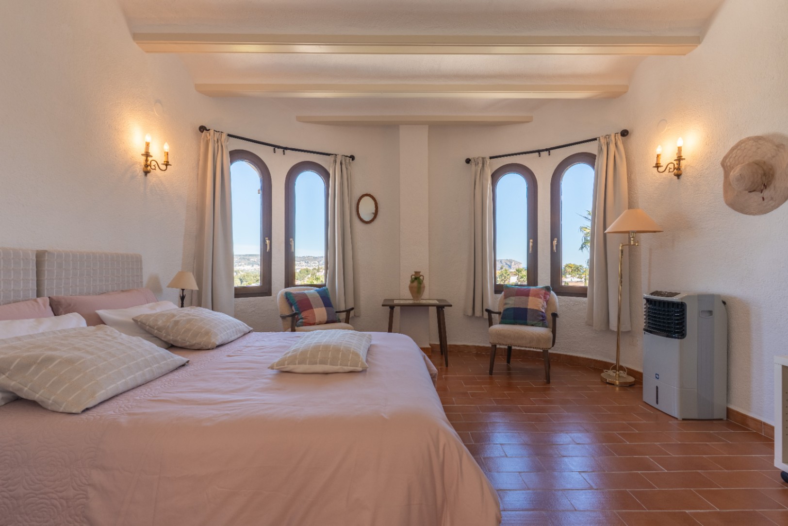 Chambre lumineuse avec sol en terre cuite, poutres apparentes et quatre fenêtres cintrées. Elle dispose d'un coin salon et d'appliques murales de style classique.
