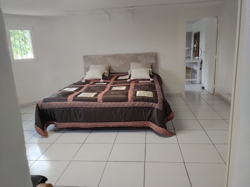 Chambre spacieuse avec sol en carrelage blanc, lit double à tête de lit fleurie et ouverture sur une pièce attenante servant de bureau ou de dressing.