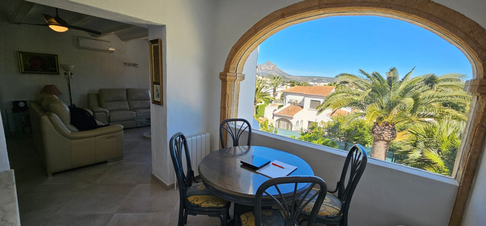 Coin repas vitré avec arche en pierre de Tosca offrant une vue sur le Montgó. L'espace communique avec un salon confortable équipé de canapés en cuir et climatisation.