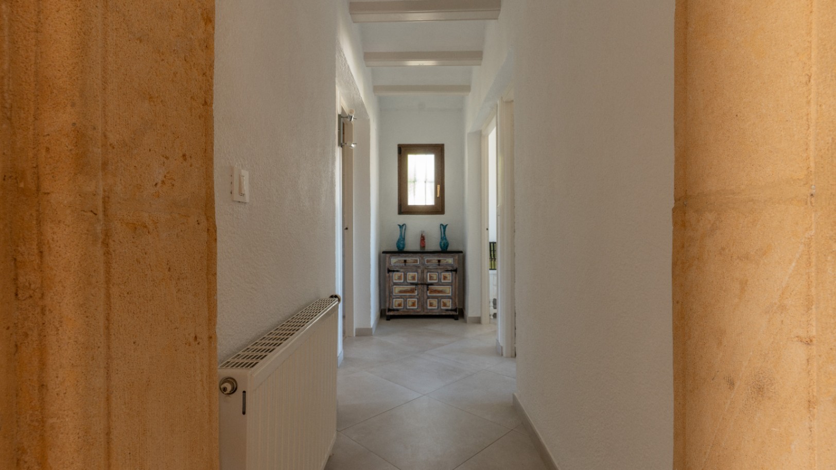 Couloir lumineux avec arches en pierre de Tosca, poutres apparentes blanches et carrelage clair. Équipé d'un radiateur et d'un buffet rustique sous une fenêtre en bois.