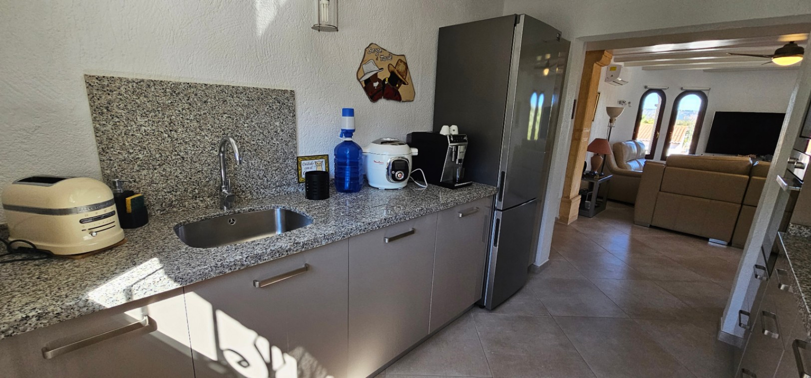 Cuisine fonctionnelle avec plan de travail en granit, évier en inox et placards modernes, s'ouvrant sur un salon lumineux aux finitions soignées.