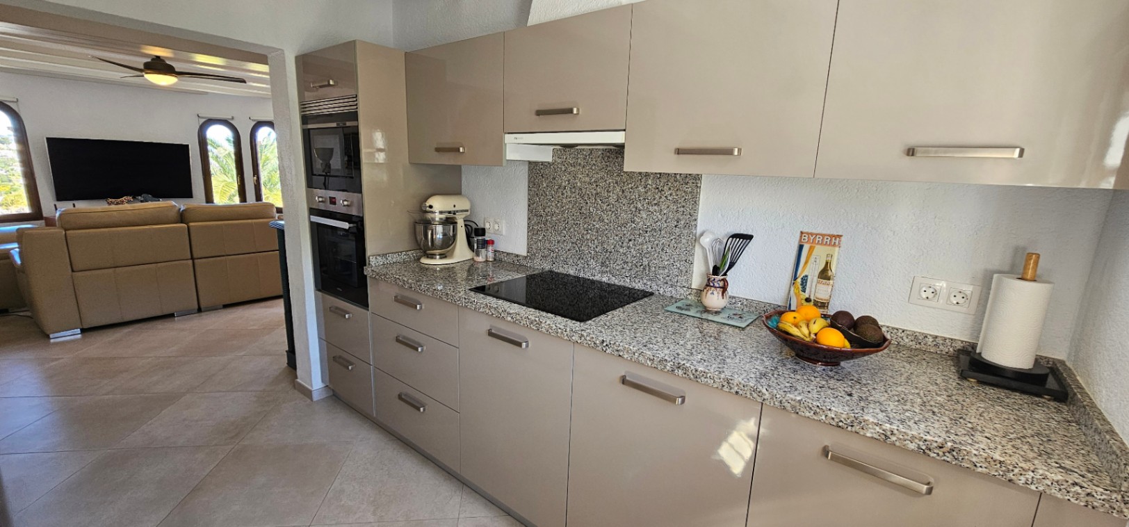 Cuisine moderne avec plans de travail en granit, meubles laqués taupe et électroménagers intégrés, s'ouvrant sur un salon lumineux aux fenêtres cintrées.