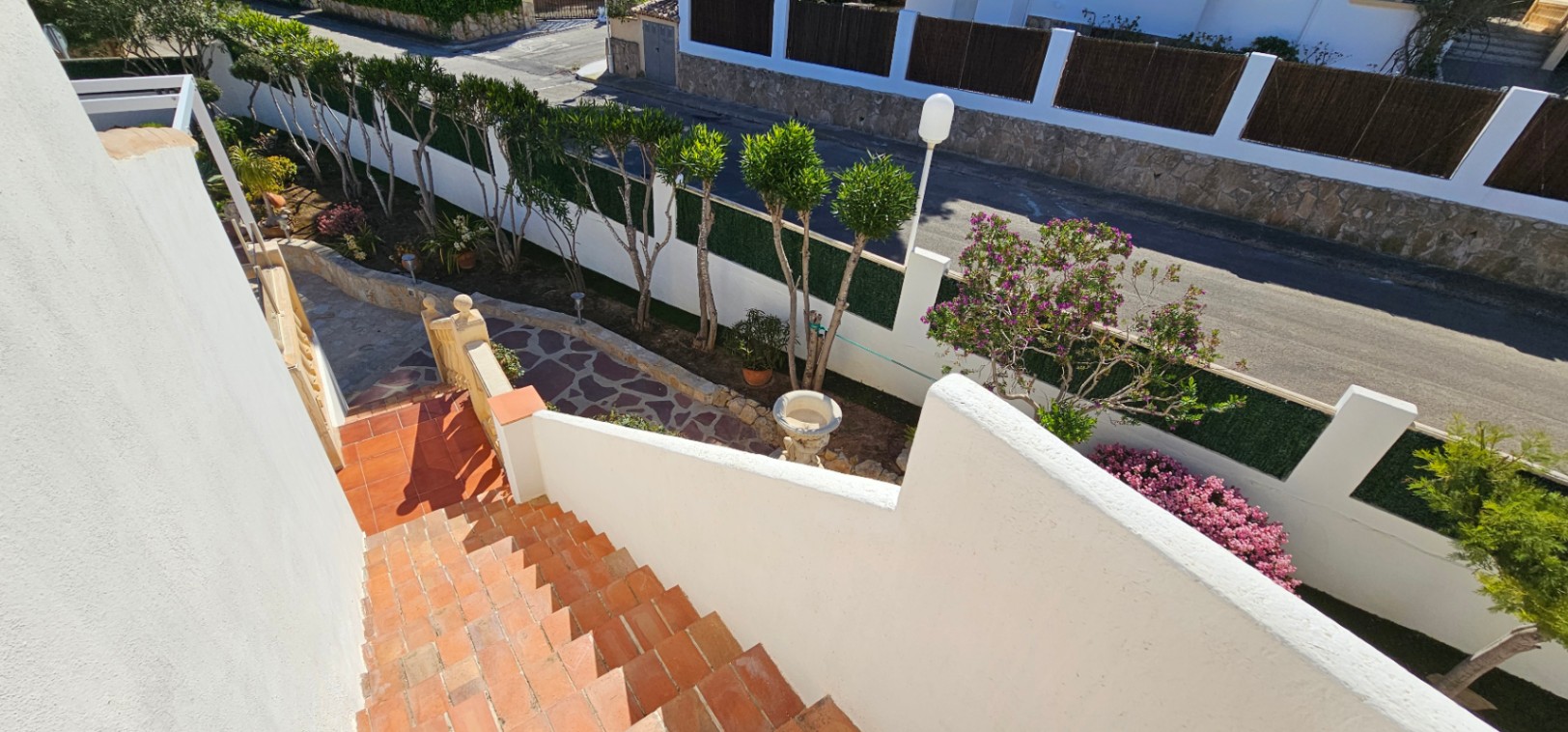 Escalier extérieur en terre cuite menant à une allée en pierre naturelle, entouré d'un jardin paysager avec des arbres ornementaux et des murs blancs.