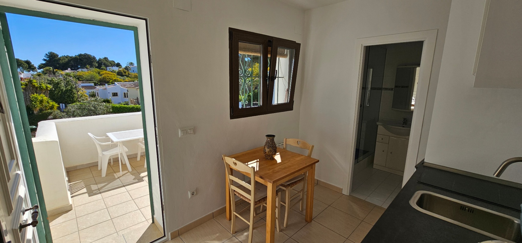 Espace repas avec table en bois, accès direct à la terrasse carrelée et vue sur la salle de bain et le plan de travail.