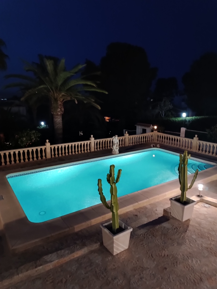 Piscine illuminée de nuit aux reflets turquoise, entourée d'une terrasse en pierre avec balustrade classique, cactus en pot et végétation méditerranéenne.
