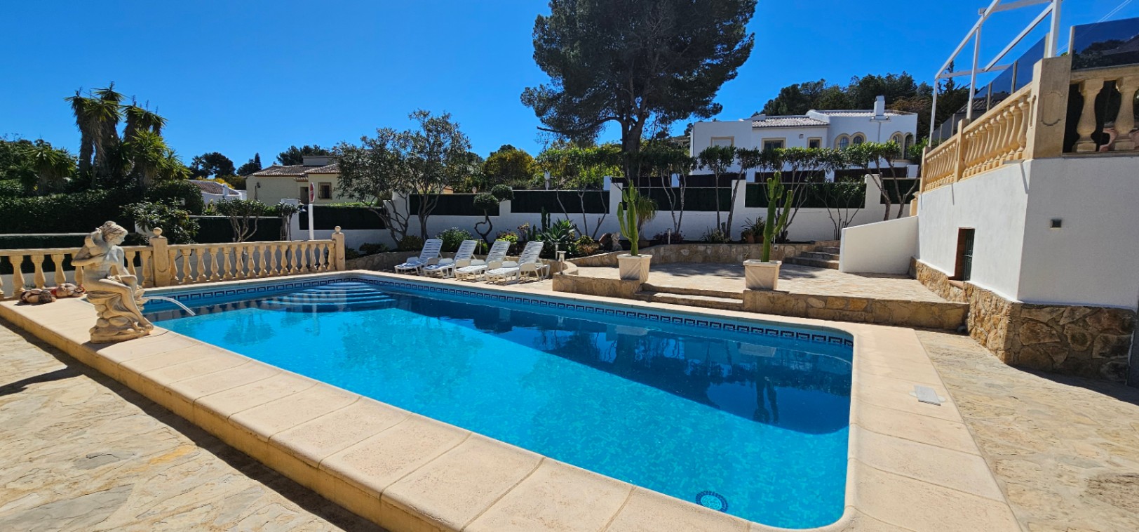 Piscine rectangulaire avec terrasse en pierre, bains de soleil blancs et balustrades classiques, entourée d'un jardin méditerranéen et de cactus.
