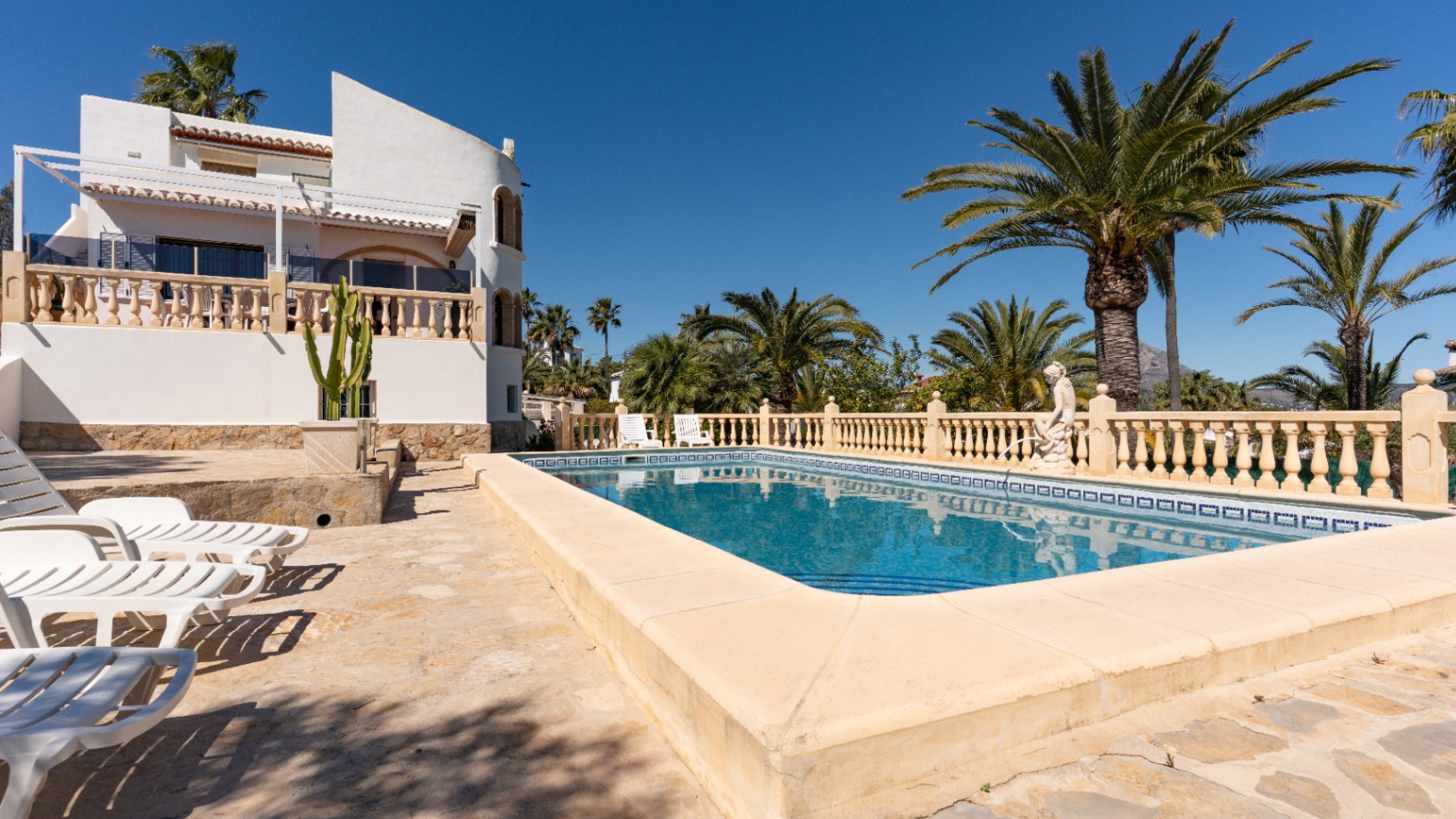 Piscine rectangulaire avec terrasse en pierre et bains de soleil, entourée de palmiers et d'une façade méditerranéenne blanche aux balustrades classiques.