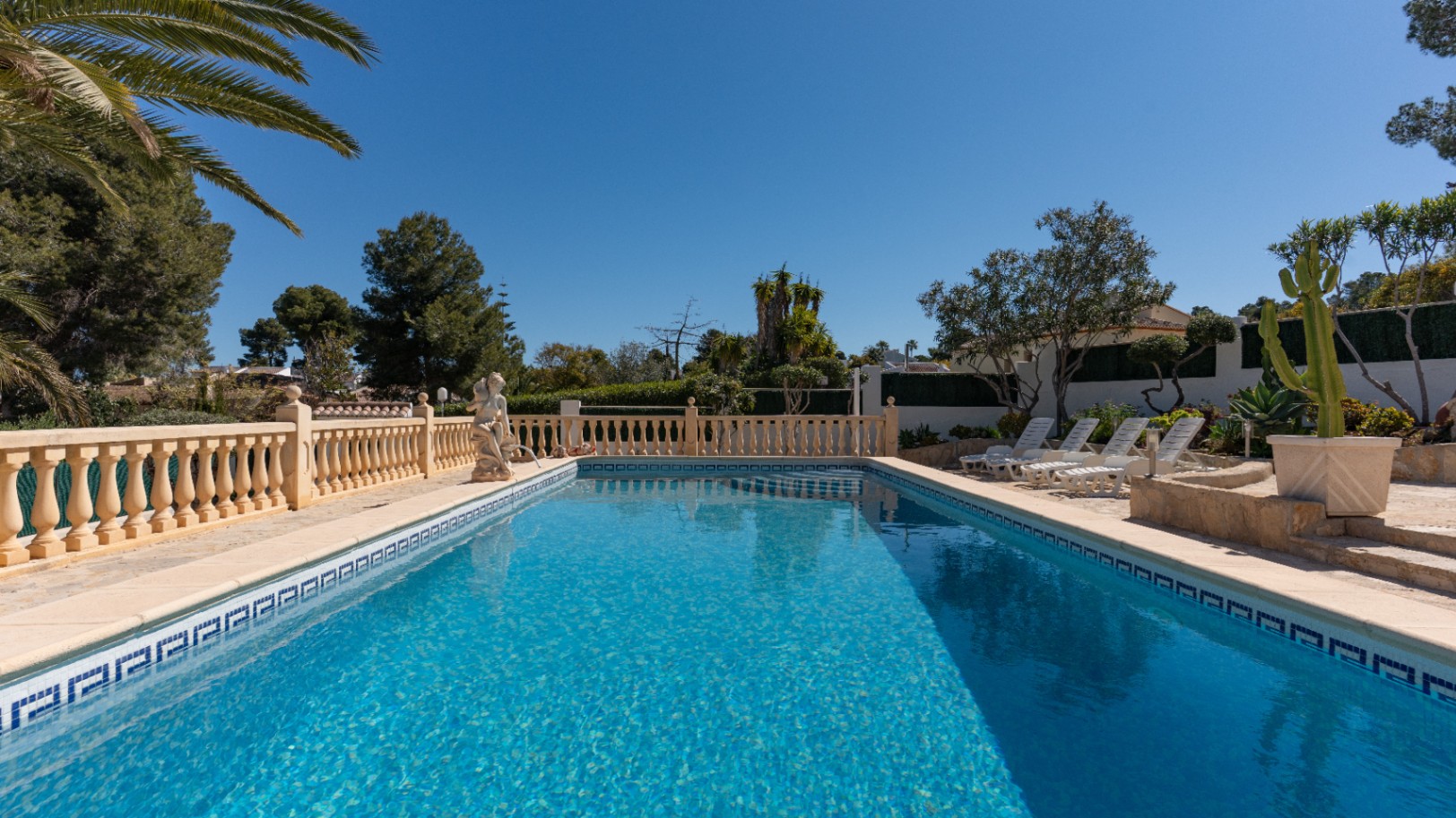 Piscine rectangulaire en mosaïque bleue entourée d'une terrasse en pierre avec balustrade classique, bains de soleil et jardin méditerranéen arboré de palmiers.