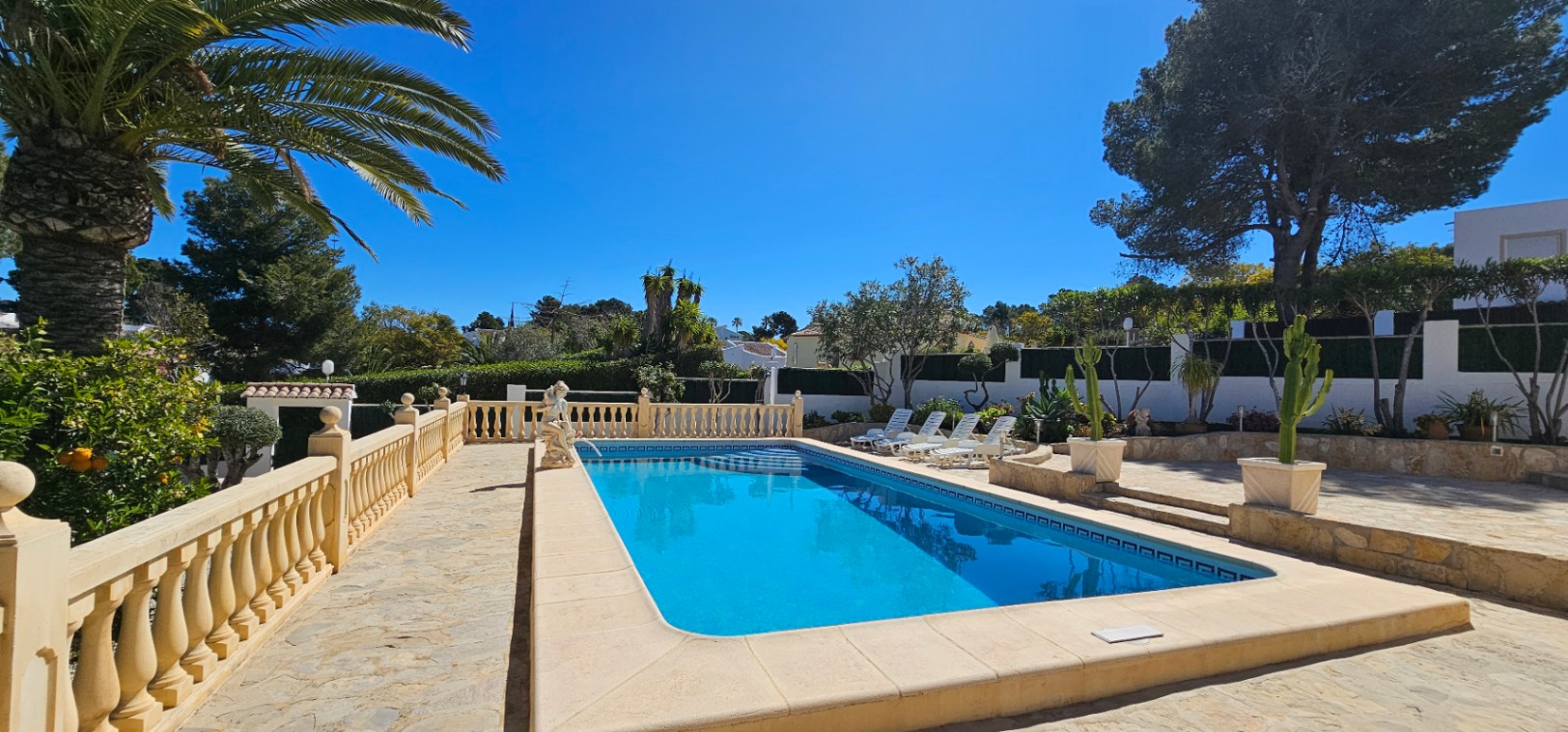 Piscine rectangulaire entourée d'une terrasse en pierre avec balustrade classique, espace de détente avec transats et jardin méditerranéen soigné.