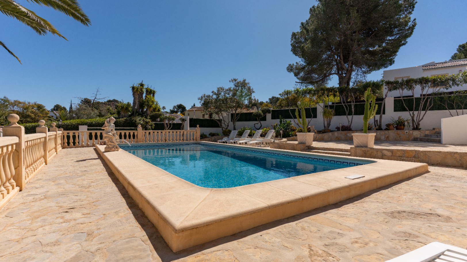 Piscine rectangulaire entourée d'une terrasse en pierre naturelle, avec balustrade classique, statue décorative et espace solarium parmi une végétation méditerranéenne.