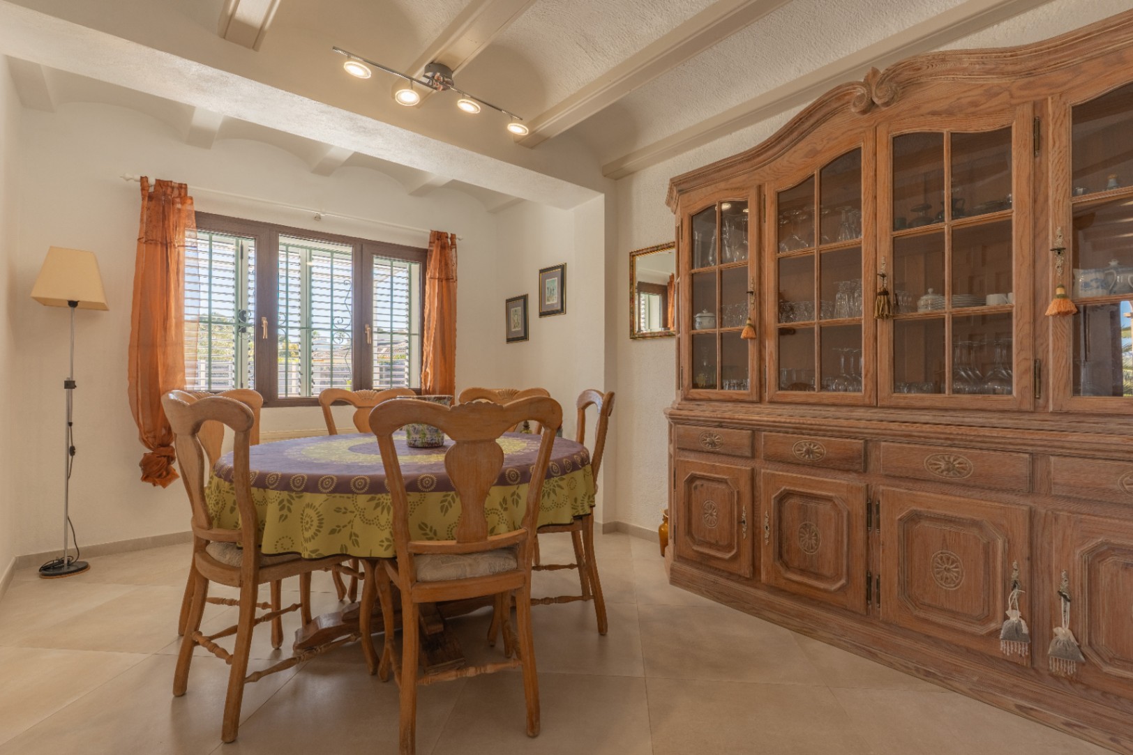 Salle à manger rustique avec poutres apparentes, table ronde en bois et grand vaisselier classique. La fenêtre à persiennes diffuse une lumière naturelle douce.