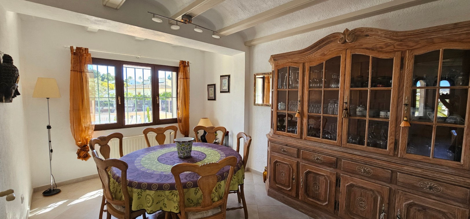 Salle à manger traditionnelle avec poutres apparentes, grand buffet en bois sculpté et table ronde. La fenêtre en bois offre une lumière naturelle et une vue sur l'extérieur.