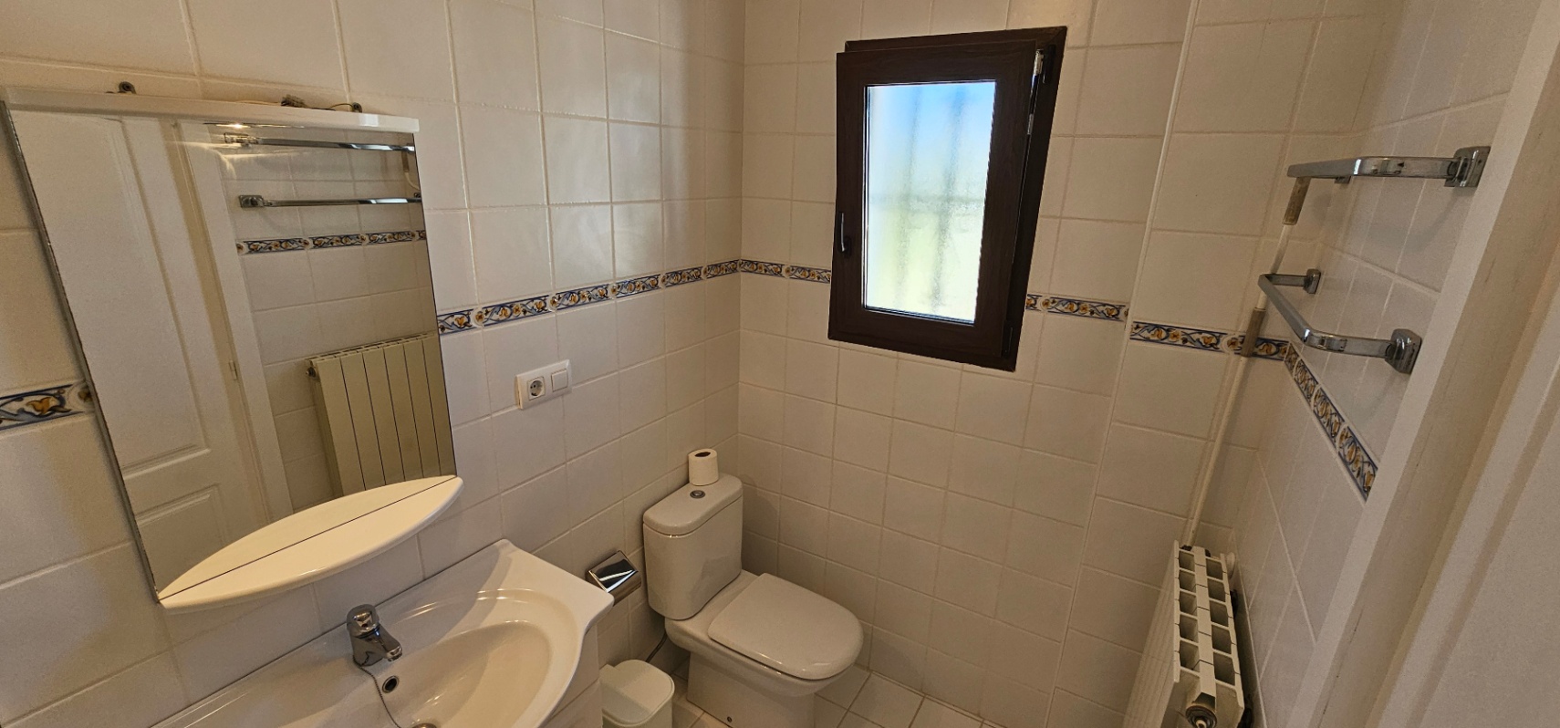 Salle de bain fonctionnelle avec carrelage blanc et frise décorative. Elle dispose d'un lavabo en céramique, d'un radiateur et d'une fenêtre givrée pour l'intimité.