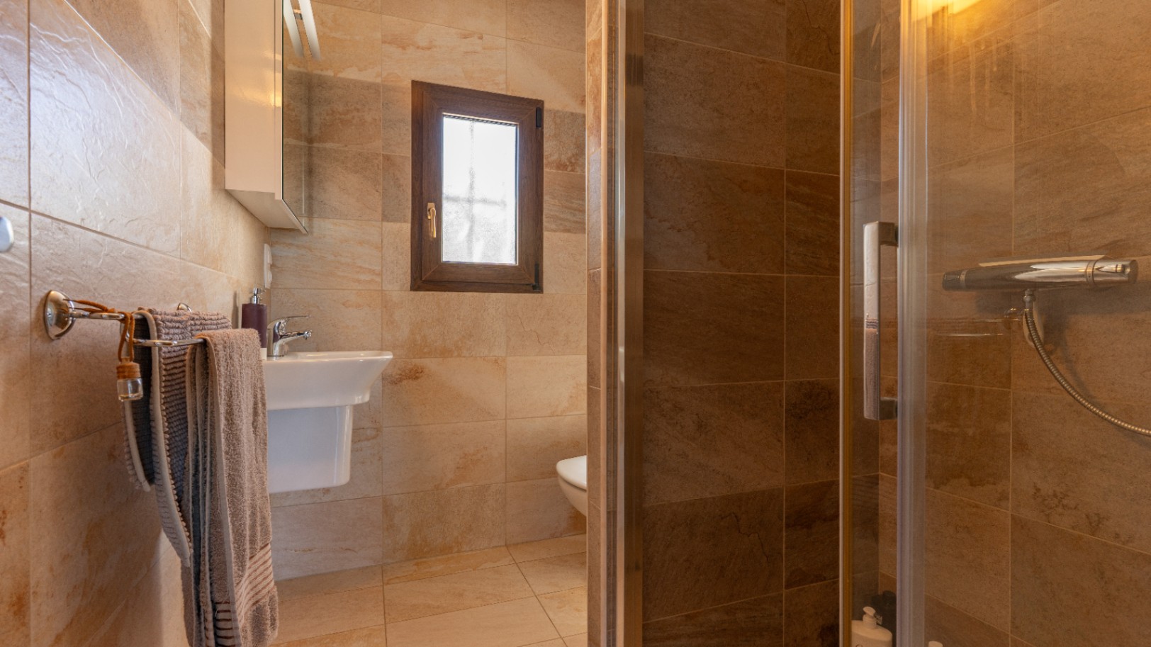 Salle de bain revêtue de carreaux céramiques aux tons terre, avec douche vitrée, lavabo suspendu et fenêtre en bois à vitrage dépoli.