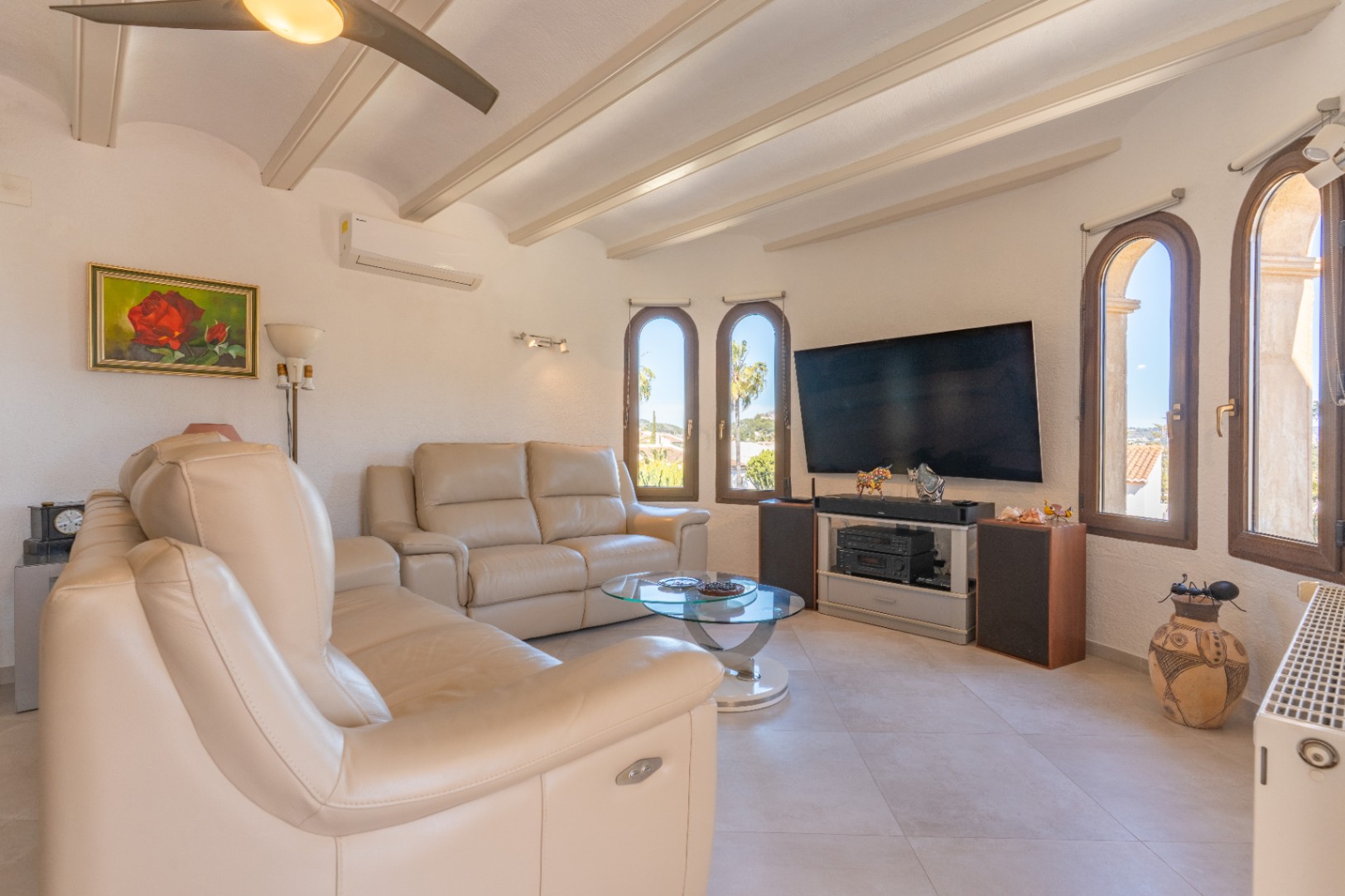 Salon lumineux avec poutres apparentes, canapés en cuir crème et fenêtres voûtées. Équipé de la climatisation, d'un ventilateur de plafond et d'un coin multimédia.