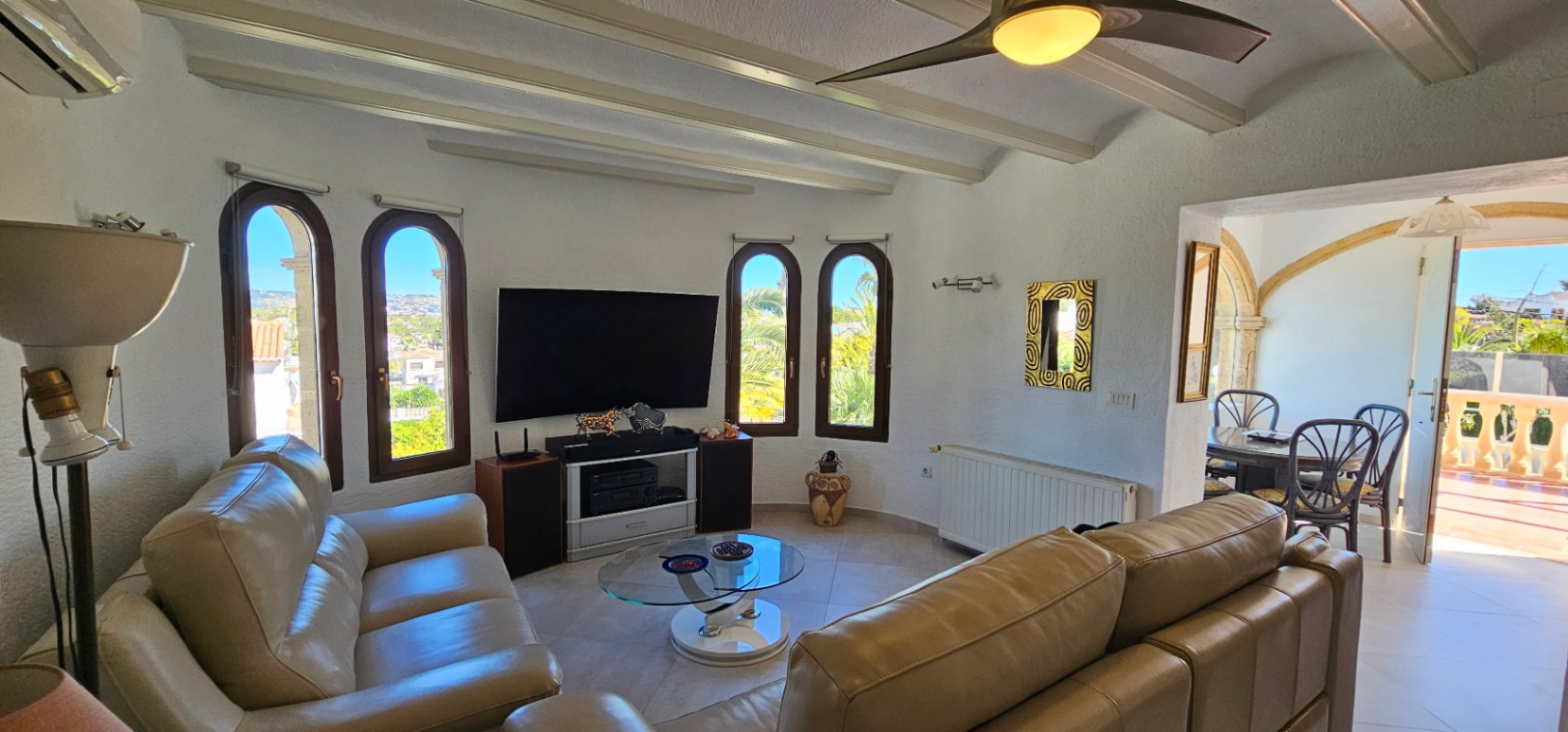Séjour avec plafonds à poutres blanches, canapés en cuir et fenêtres en arc. L'espace communique par une arche en pierre avec la salle à manger et la terrasse.