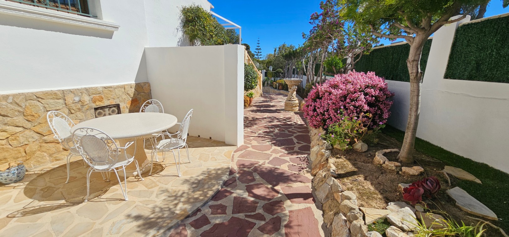 Terrasse extérieure avec sol en pierre naturelle, salon de jardin en fer forgé blanc et jardins paysagers agrémentés de buissons fleuris et murets.