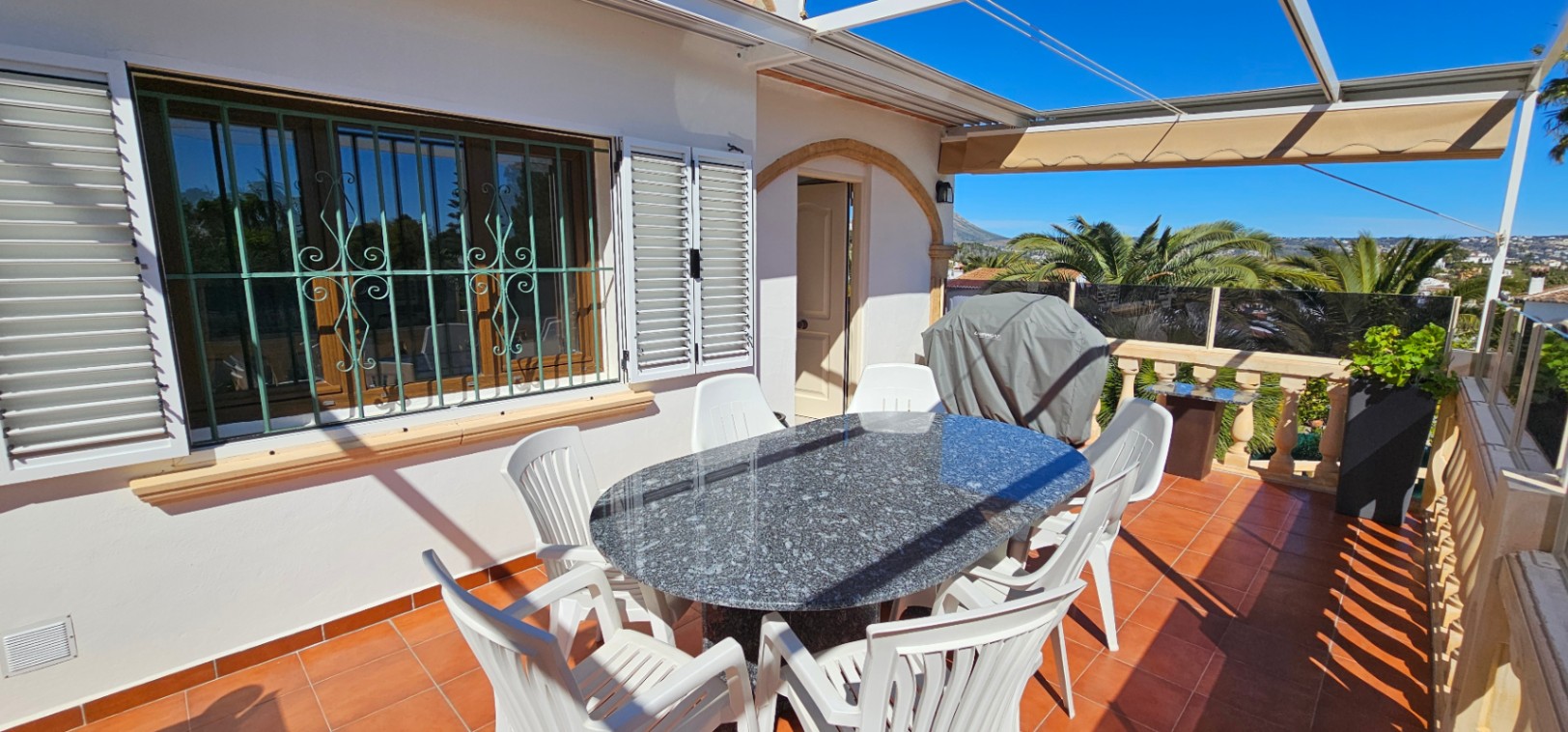 Terrasse surélevée avec sol en terre cuite, table en granit et vue dégagée. Comprend un store banne, une balustrade en pierre et des fenêtres avec volets traditionnels.