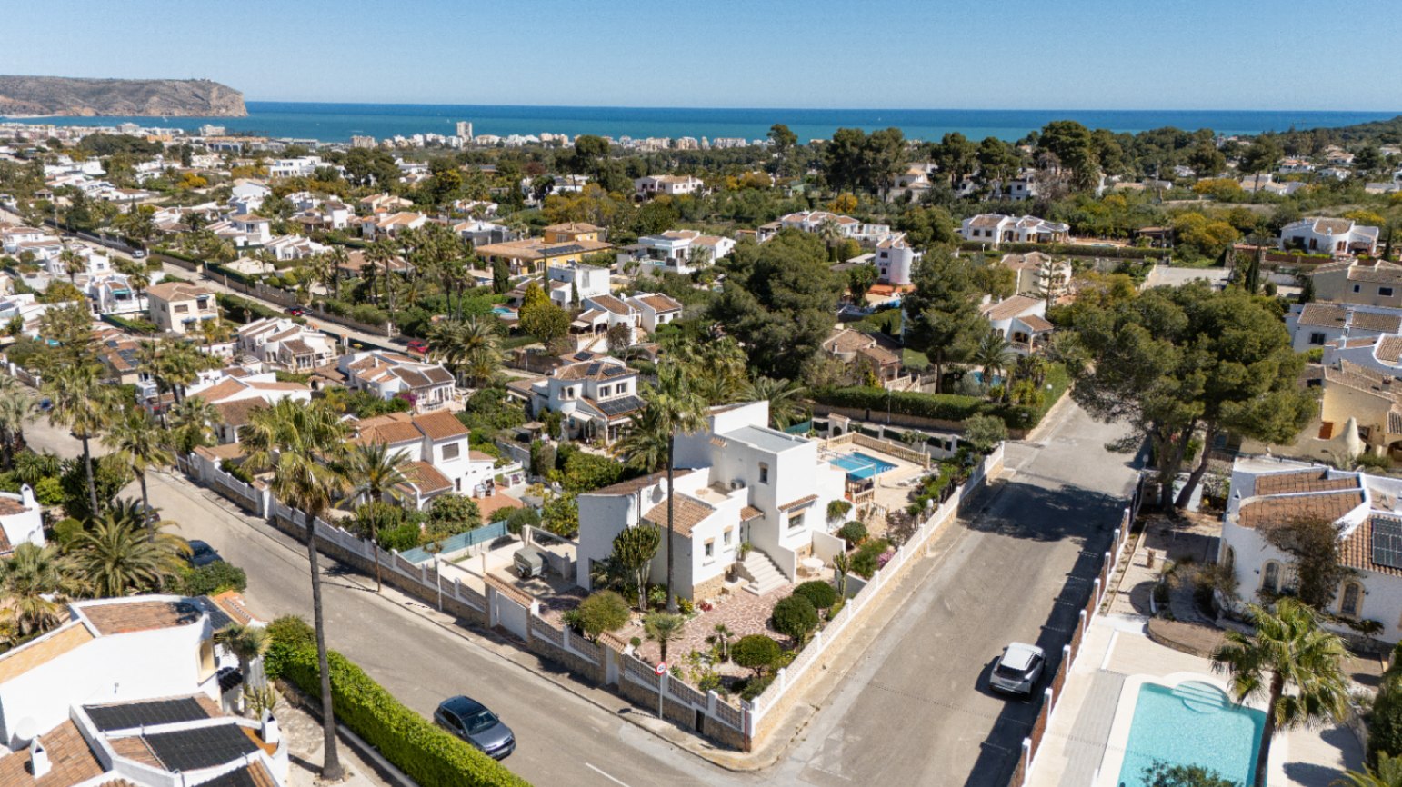 Vue aérienne de la villa blanche sur une parcelle d'angle à Pinosol, Jávea. On distingue la piscine privée, le jardin arboré et une vue dégagée sur la mer Méditerranée au loin.