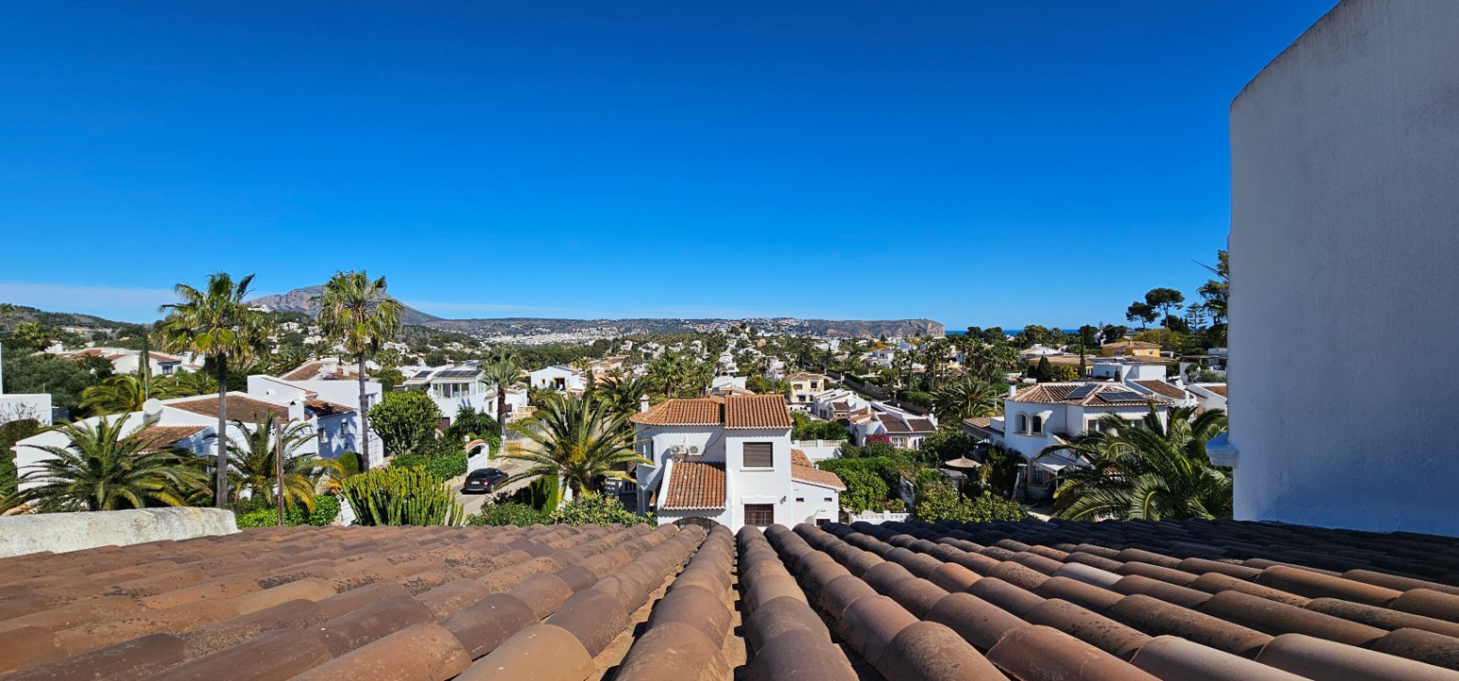 Vue panoramique depuis le toit en tuiles traditionnelles sur le quartier résidentiel de Jávea, avec ses palmiers et les montagnes à l'horizon.