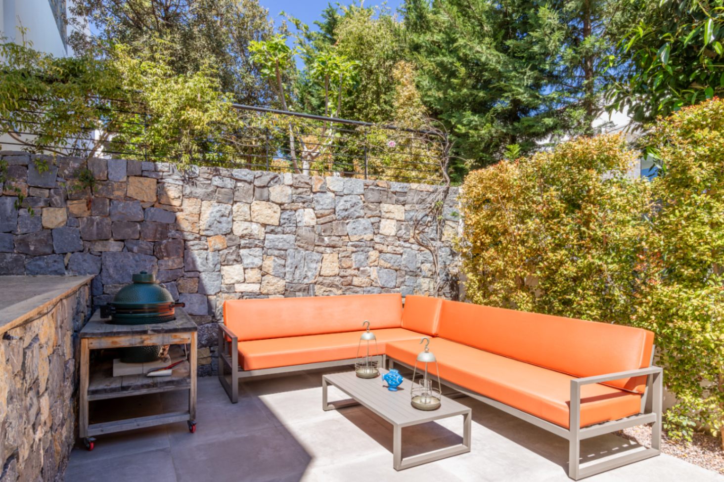 Espace de détente extérieur avec canapé d'angle orange, barbecue en céramique et mur en pierre naturelle entouré d'une végétation dense.