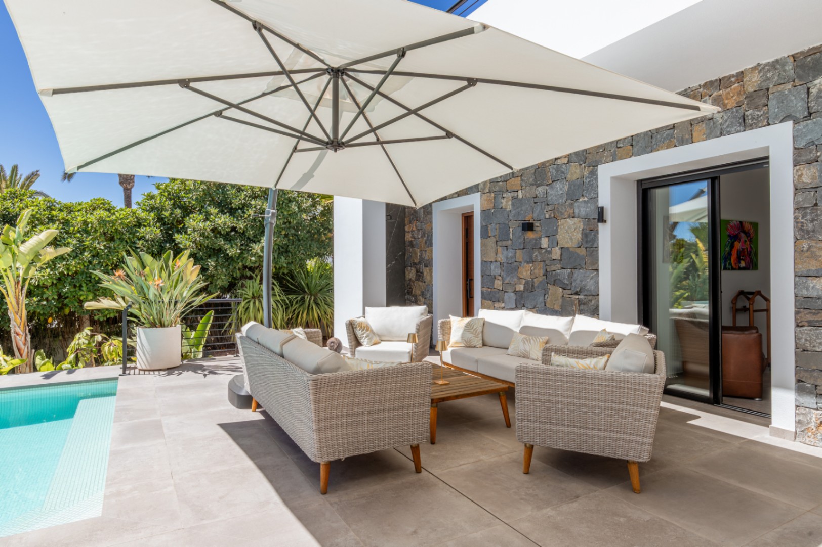 Espace de détente extérieur avec mobilier en osier et grand parasol, situé sur une terrasse carrelée bordant la piscine et un mur en pierre naturelle.