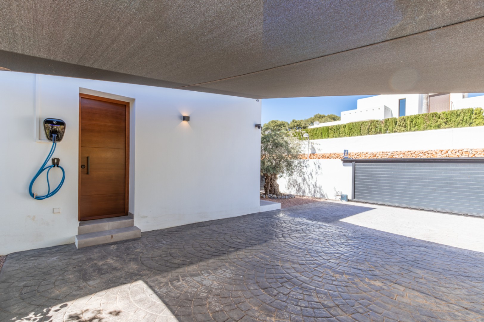 Espace de parking couvert par une voile d'ombrage, incluant une borne de recharge pour véhicules électriques et un accès direct à la maison par une porte en bois.