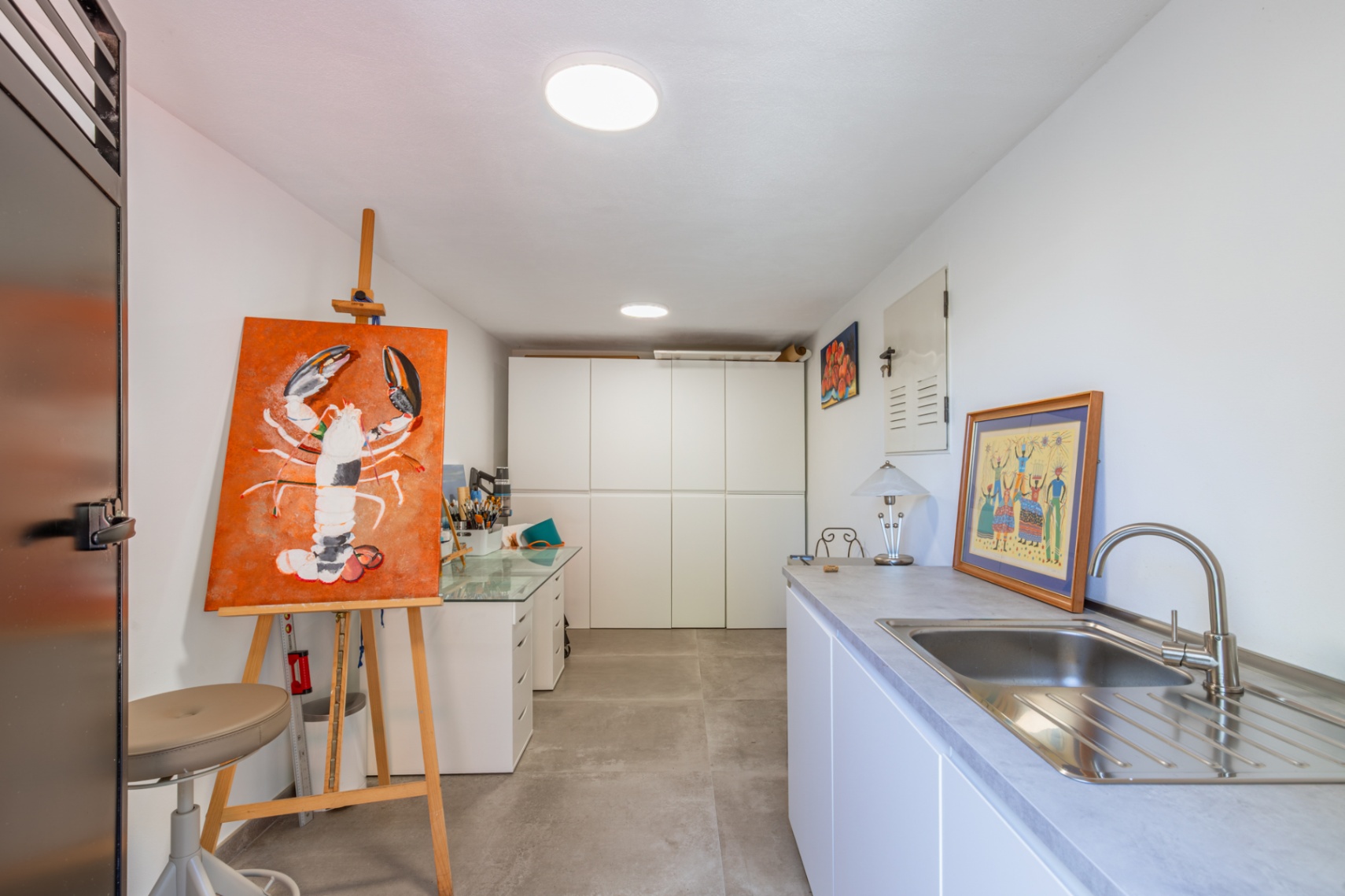 Espace polyvalent servant d'atelier ou de buanderie, équipé de rangements blancs, d'un évier en inox et d'un sol en grès cérame gris.