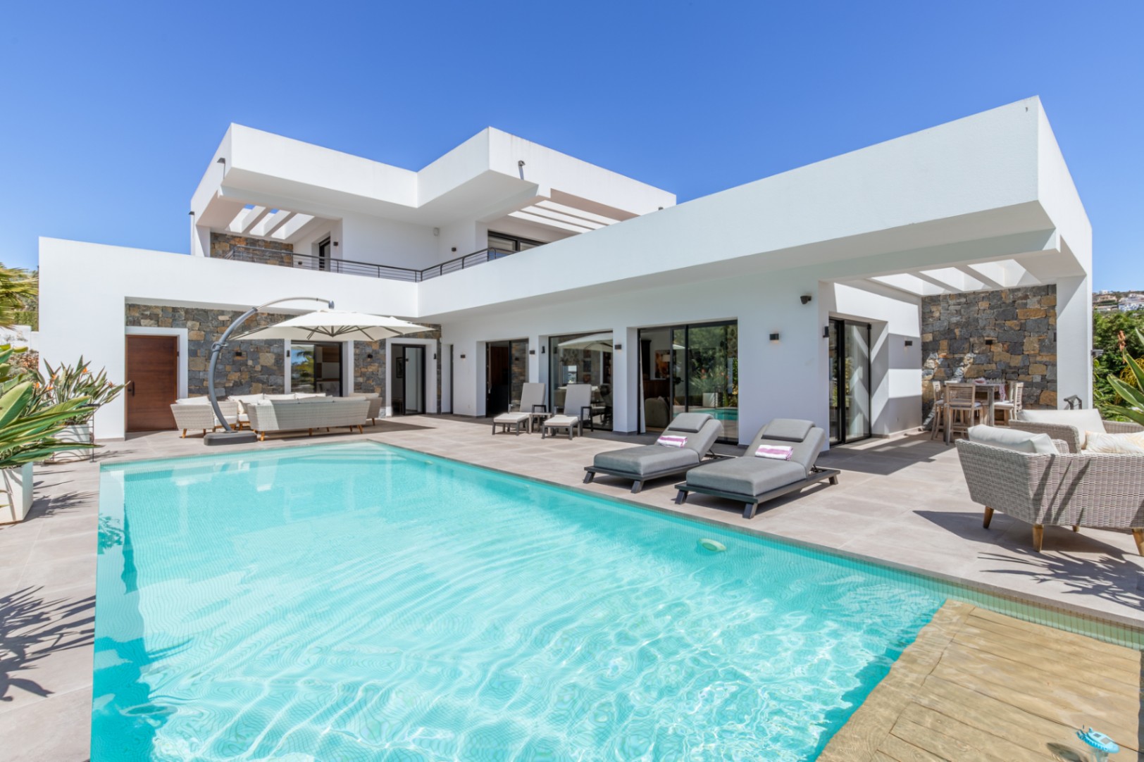 Extérieur de la villa avec piscine, terrasse spacieuse et façade blanche moderne intégrant des murs en pierre naturelle et des lignes épurées.