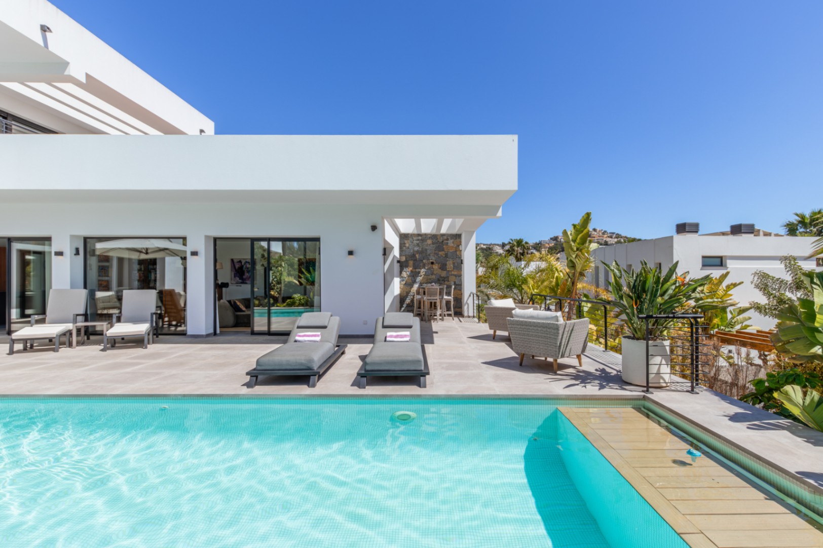 Grande terrasse avec piscine en mosaïque turquoise, espace solarium et coin repas extérieur sous pergola, intégrés dans une architecture moderne aux lignes épurées.