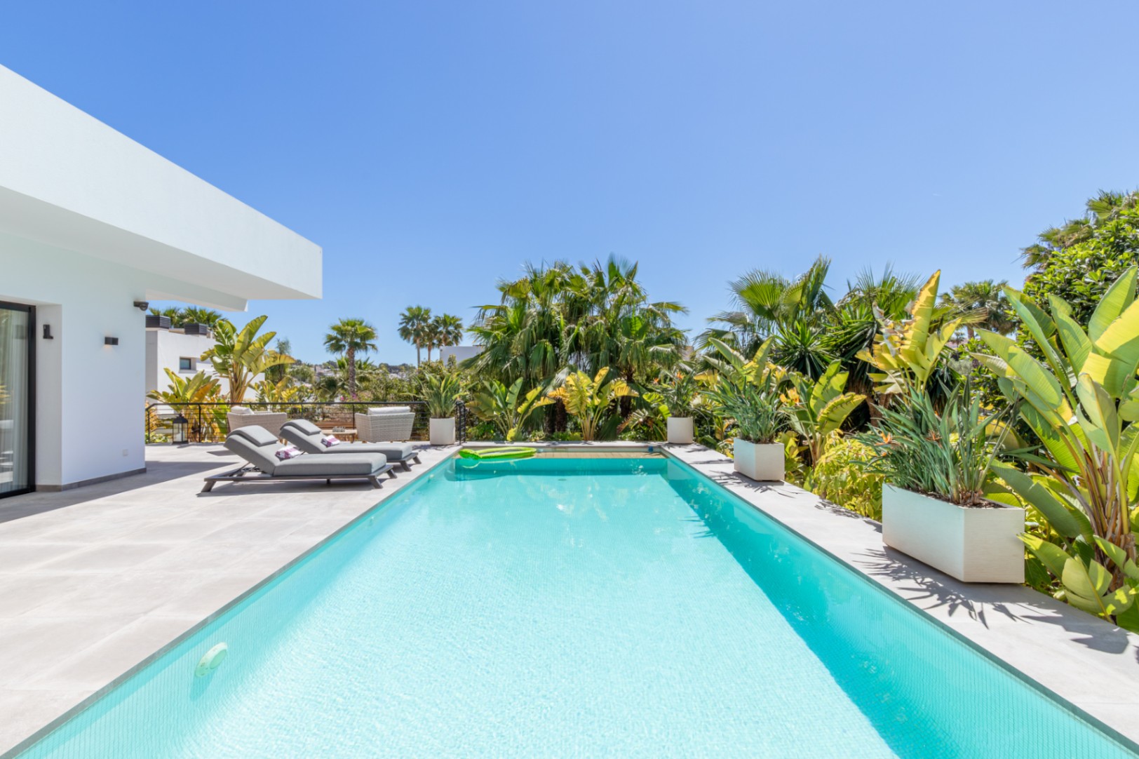 Piscine rectangulaire avec terrasse en grès cérame gris, entourée d'une végétation tropicale luxuriante et de bains de soleil modernes sous un ciel azur.