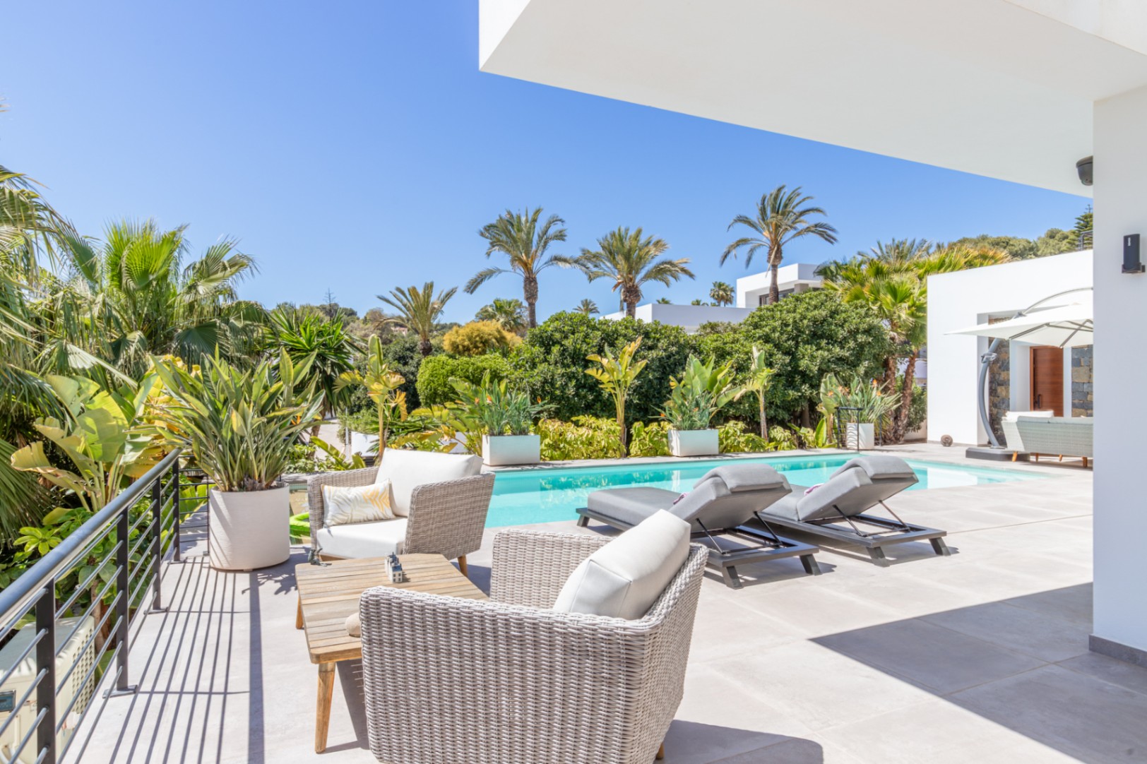 Terrasse extérieure avec mobilier en rotin et bains de soleil face à la piscine, entourée d'un jardin méditerranéen mûr avec palmiers et verdure.