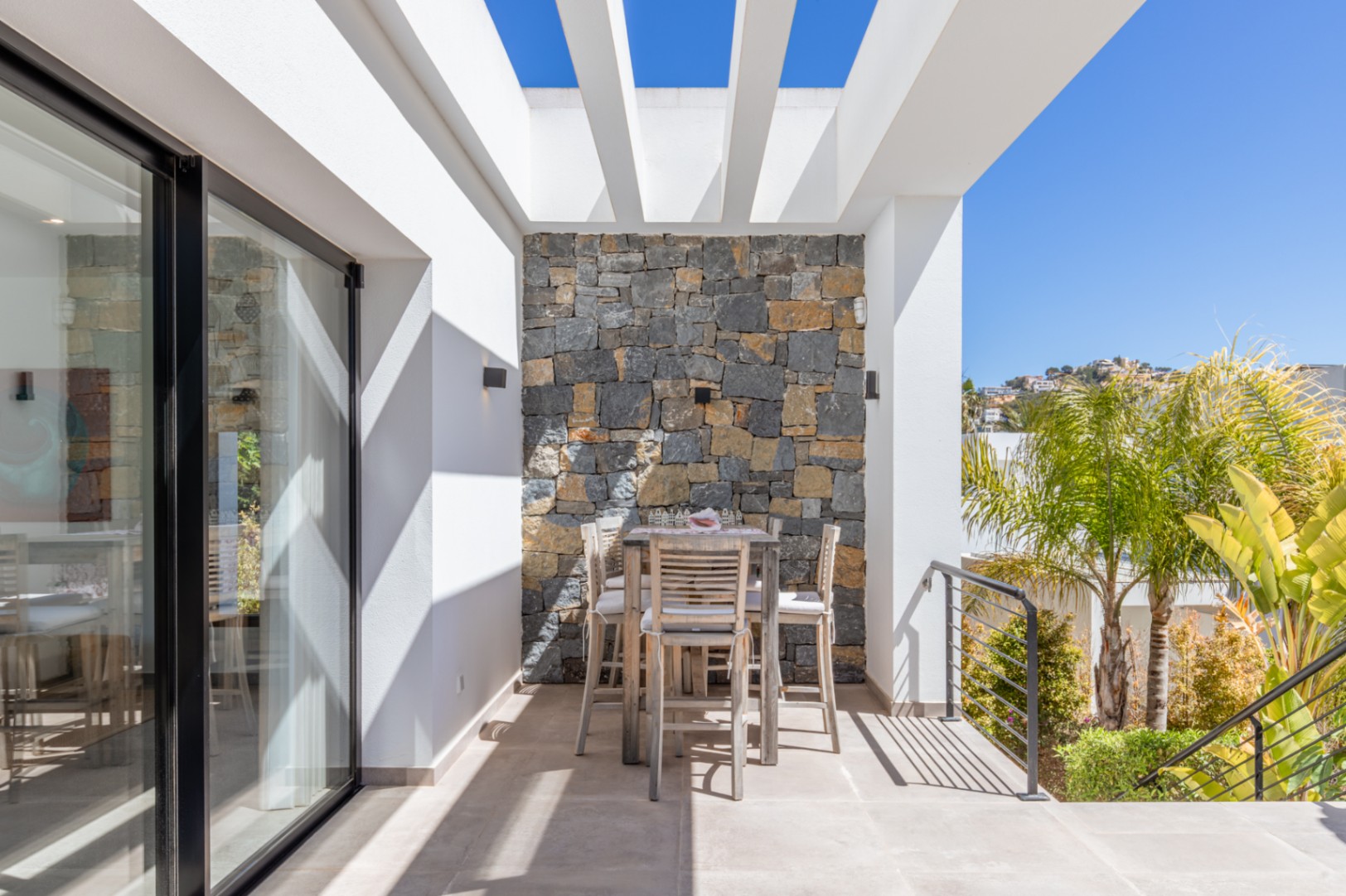 Terrasse extérieure avec pergola blanche, mur en pierre naturelle et coin repas surélevé en bois. Les grandes baies vitrées assurent une transition fluide vers l'intérieur.