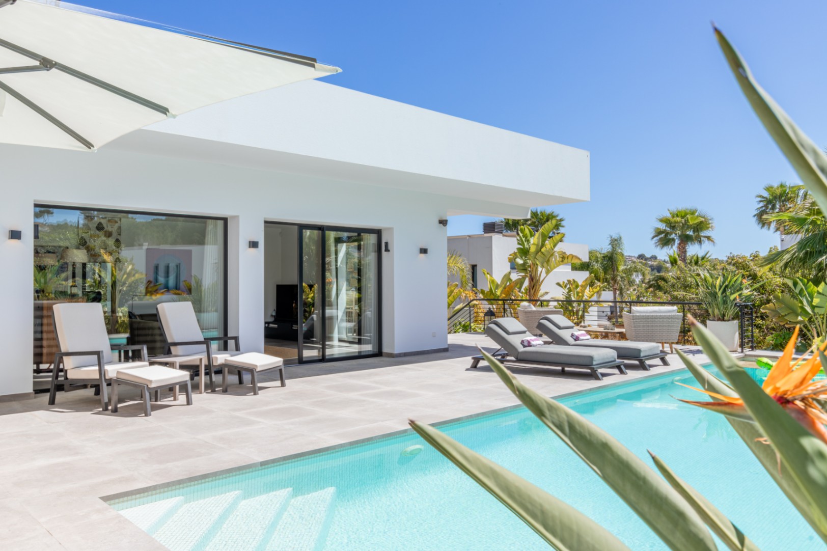 Terrasse extérieure avec piscine, bains de soleil élégants et accès direct au salon via de larges baies vitrées noires intégrées à une façade blanche épurée.