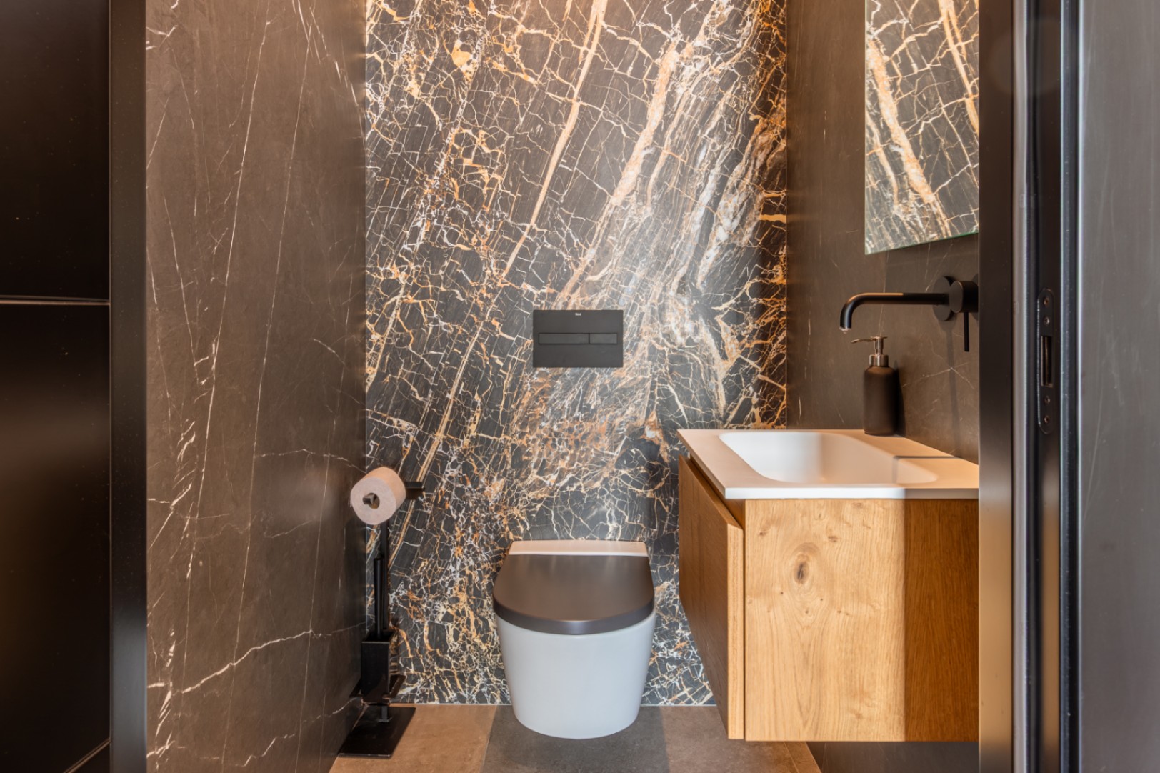 Toilettes invités avec un mur en marbre sombre aux veines dorées, un WC suspendu et un meuble sous-vasque en chêne avec robinetterie noire mate.
