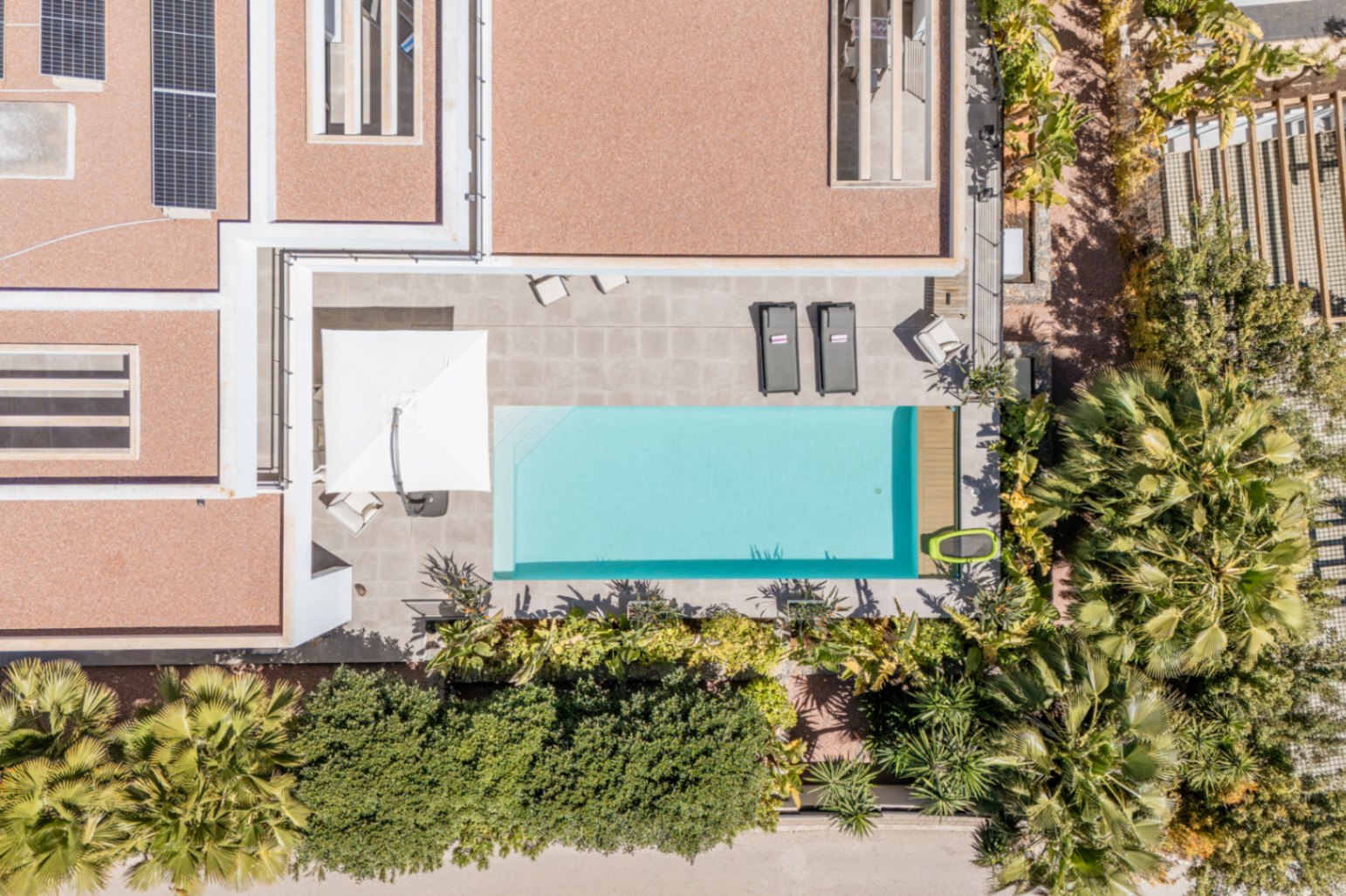 Vue aérienne de la piscine rectangulaire et de sa terrasse en grès, entourées d'une végétation méditerranéenne et de panneaux solaires en toiture.